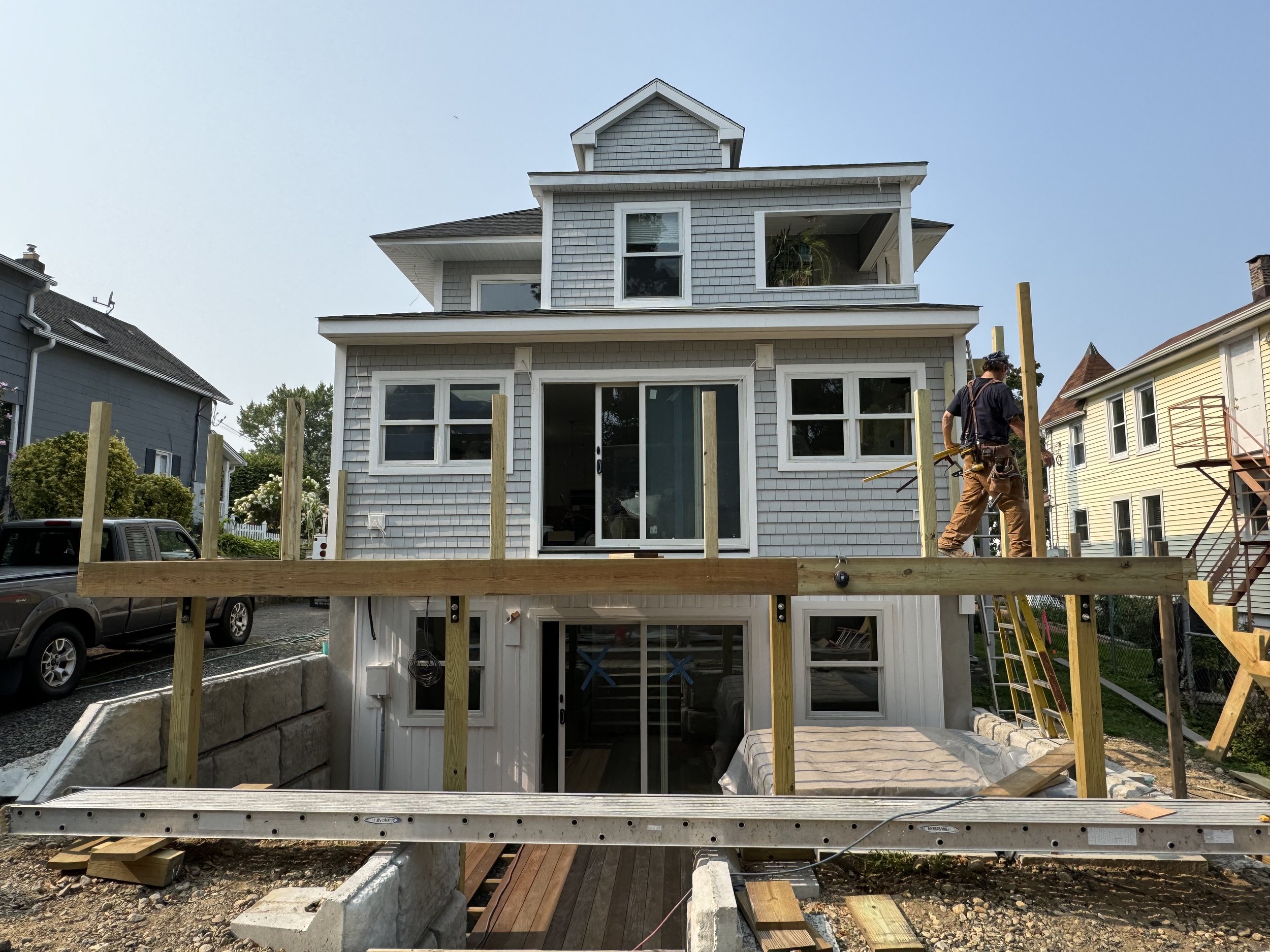 A two-story house under construction with wooden stakes set up around the perimeter, a worker installing framing, and neighboring houses visible in the background.