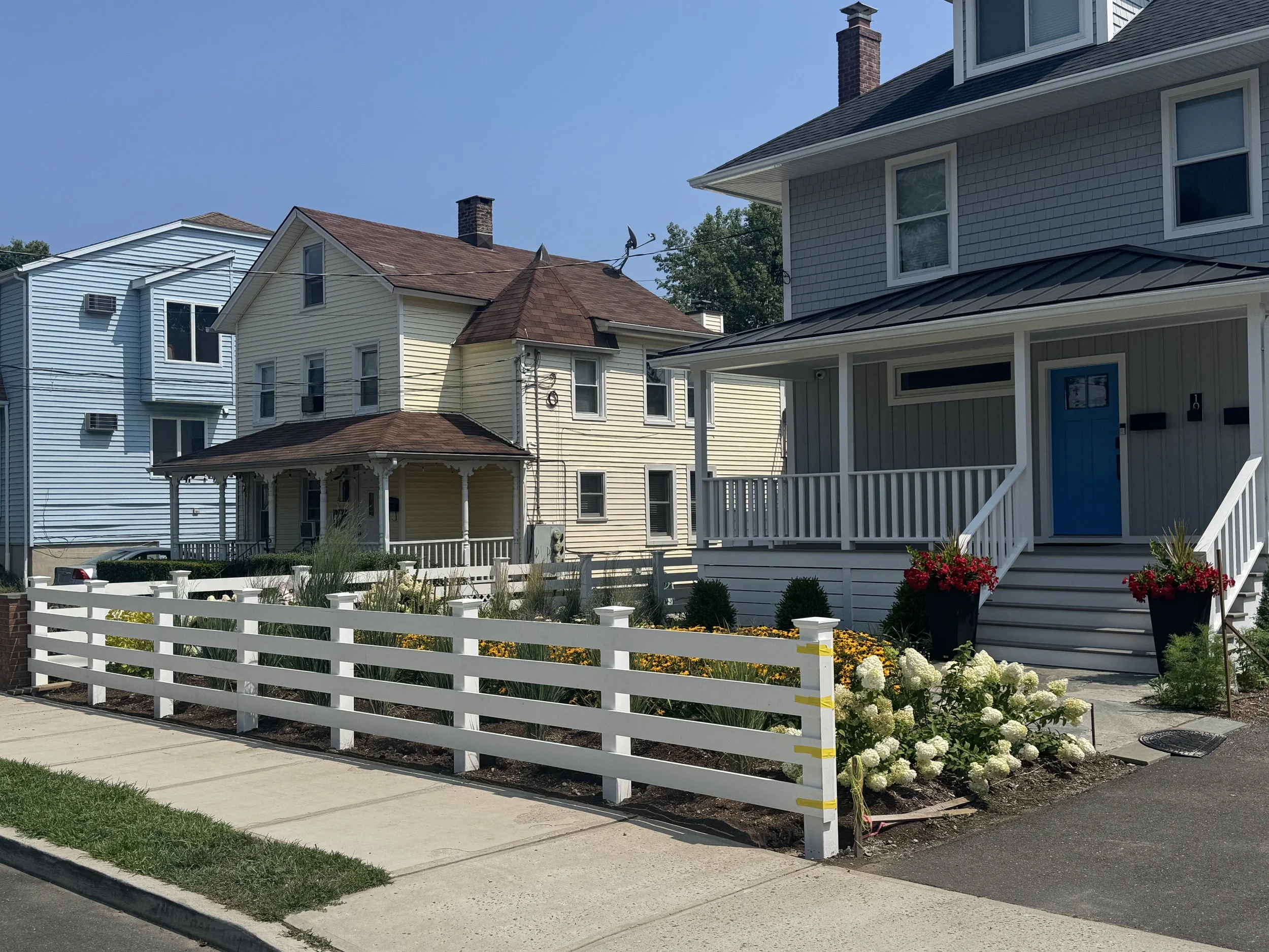 A row of colorful houses with a garden in front, surrounded by a white picket fence, along a sidewalk under a clear blue sky.