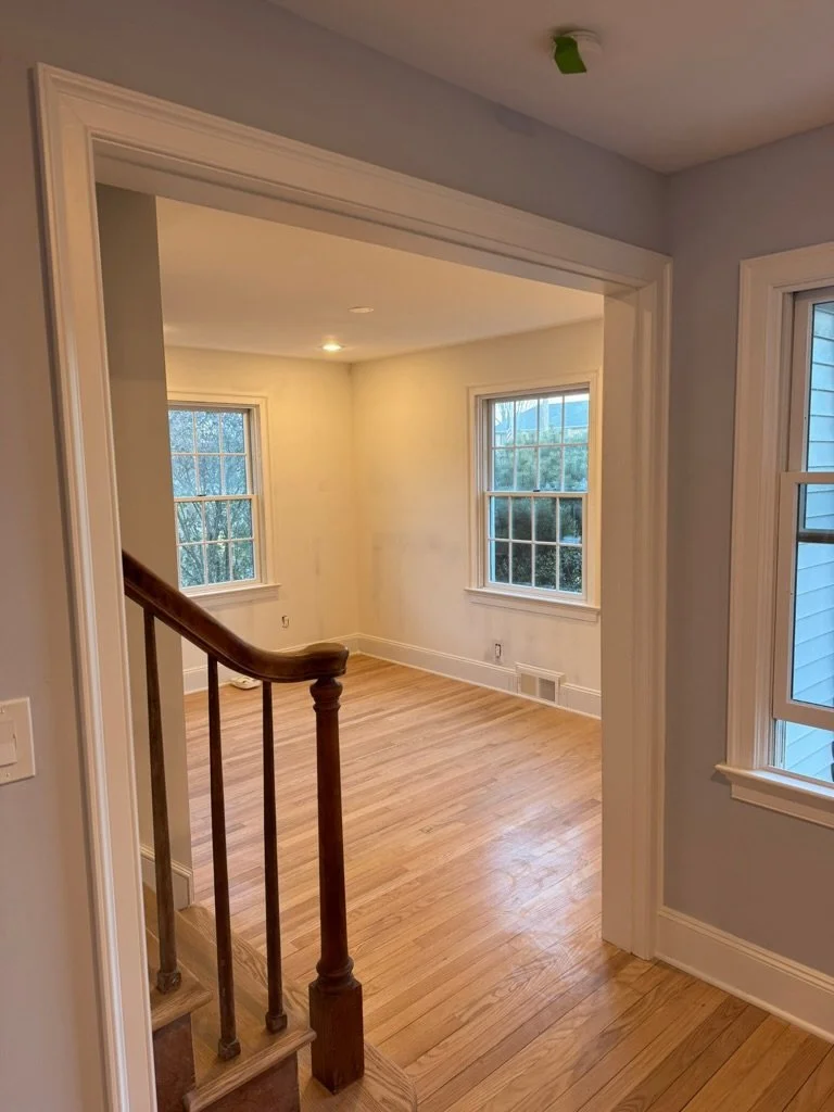 Empty room with hardwood floors, two large windows, and a staircase with wooden handrail leading into the room. Light gray walls and white trim, with natural light coming through the windows.