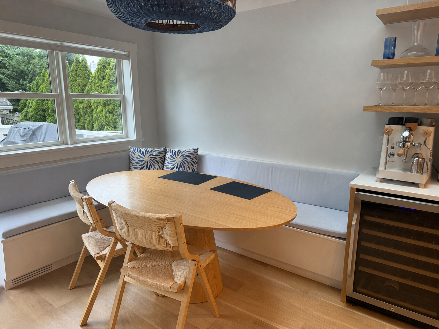 Dining nook with a round wooden table, a built-in white bench with cushions, two chairs, large window with a view of green trees, a beverage station with wine glasses, a coffee machine, and a wine cooler.
