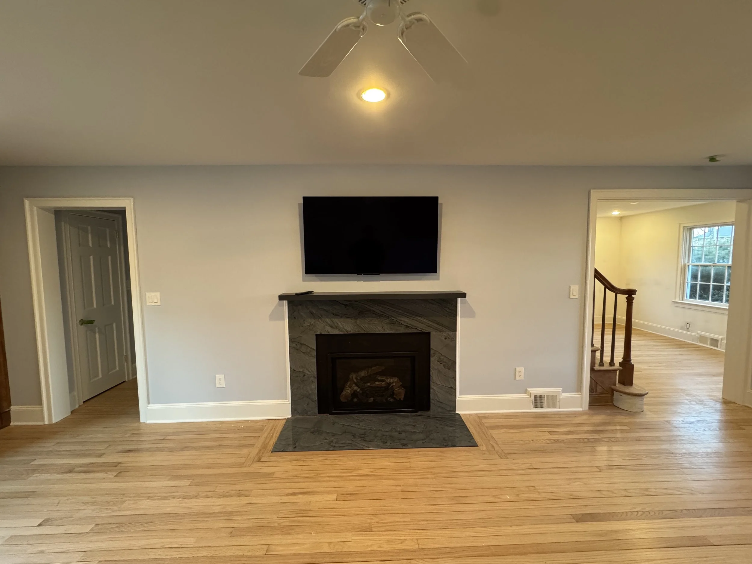 A living room with hardwood floors, a wall-mounted flat-screen TV, a fireplace with a marble surround, and a doorway leading to a staircase and another room with large windows.