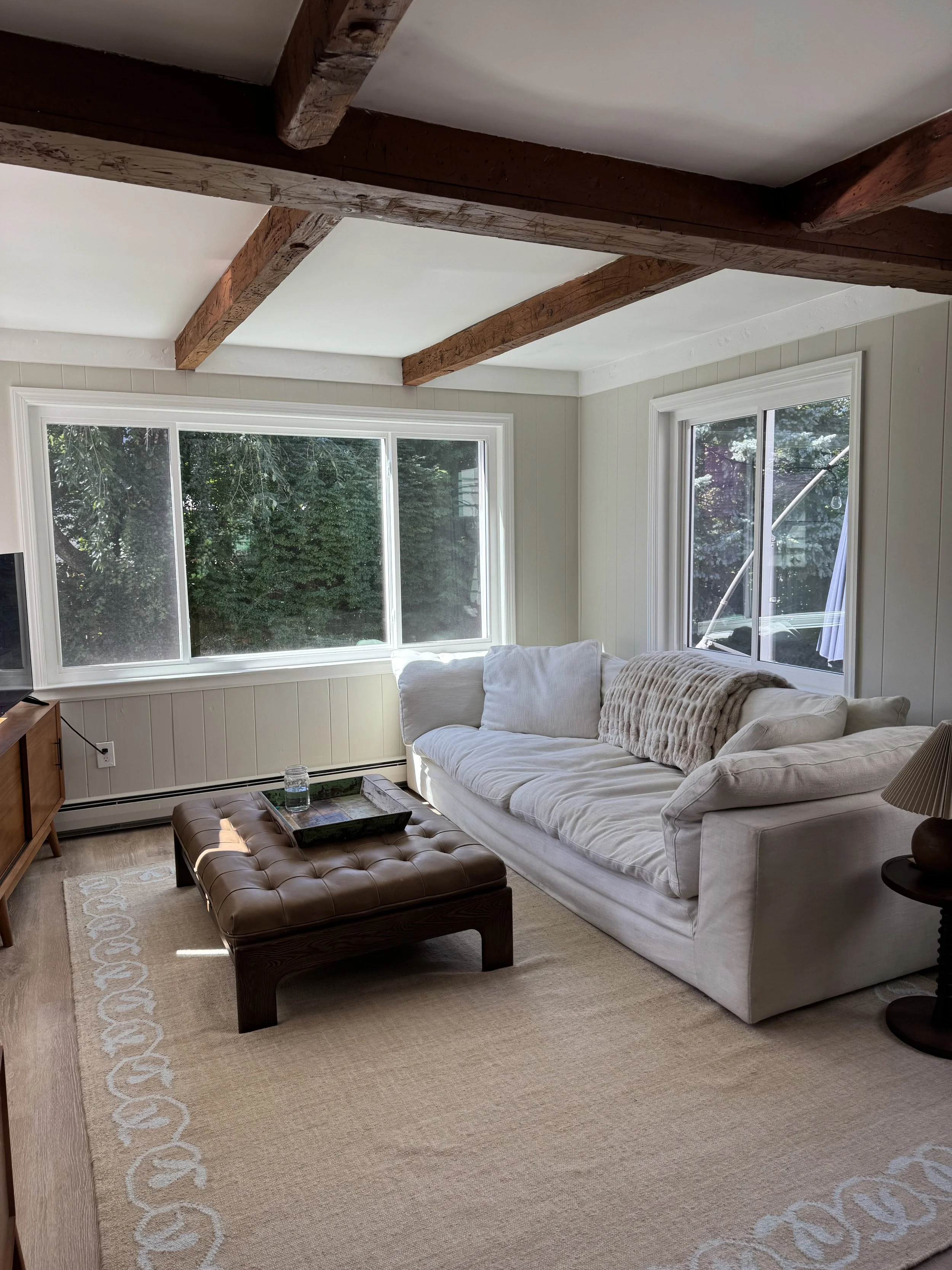 Living room with large beige area rug, white sofa with beige blanket, tufted brown ottoman, wooden side table with lamp, large windows showing trees outside, and exposed wooden beams on the ceiling.