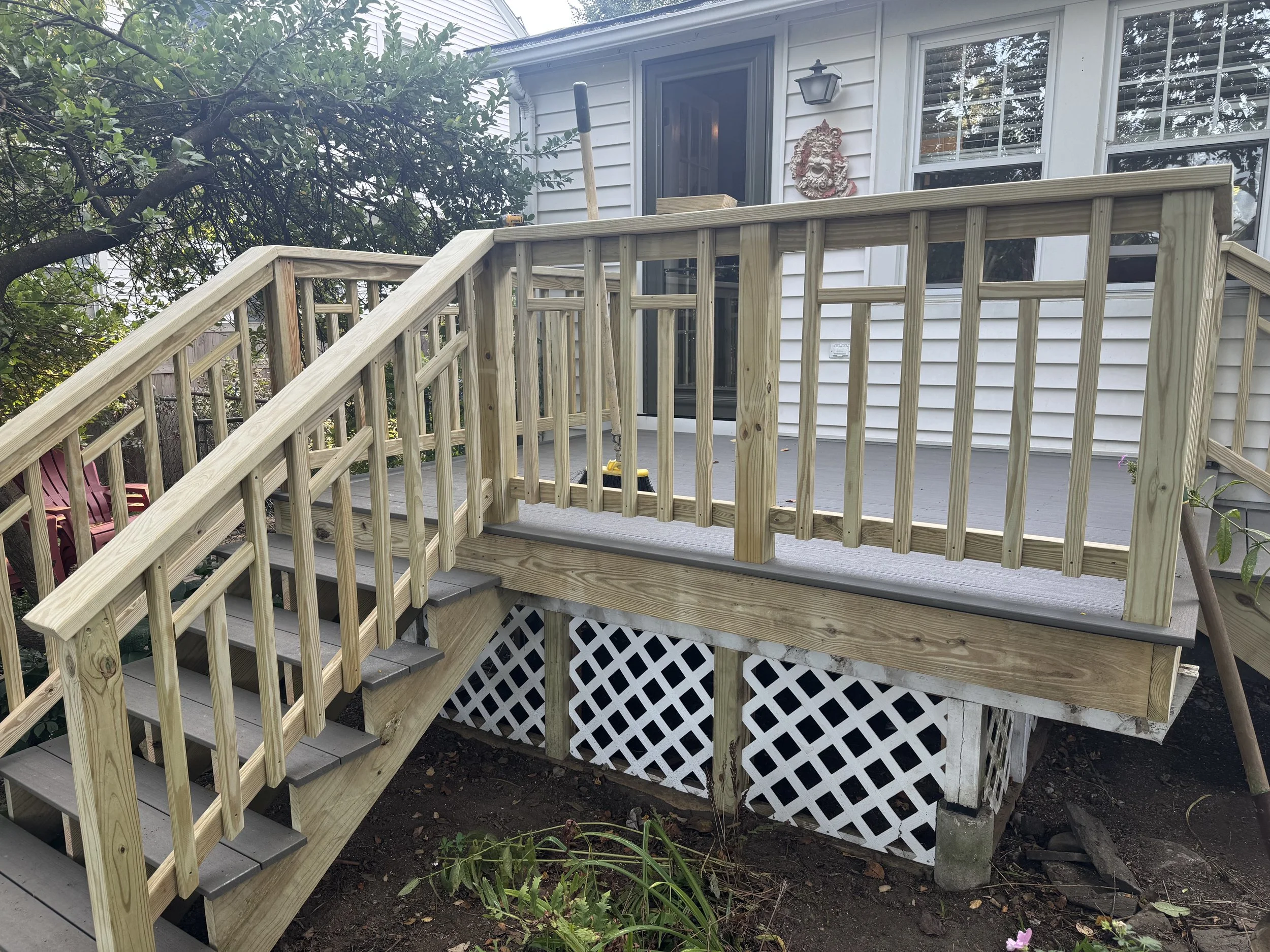 Wooden deck stairs leading to a screened door and window on a house exterior, with a decorative mask on the wall, a porch light, and a large tree on the left side.