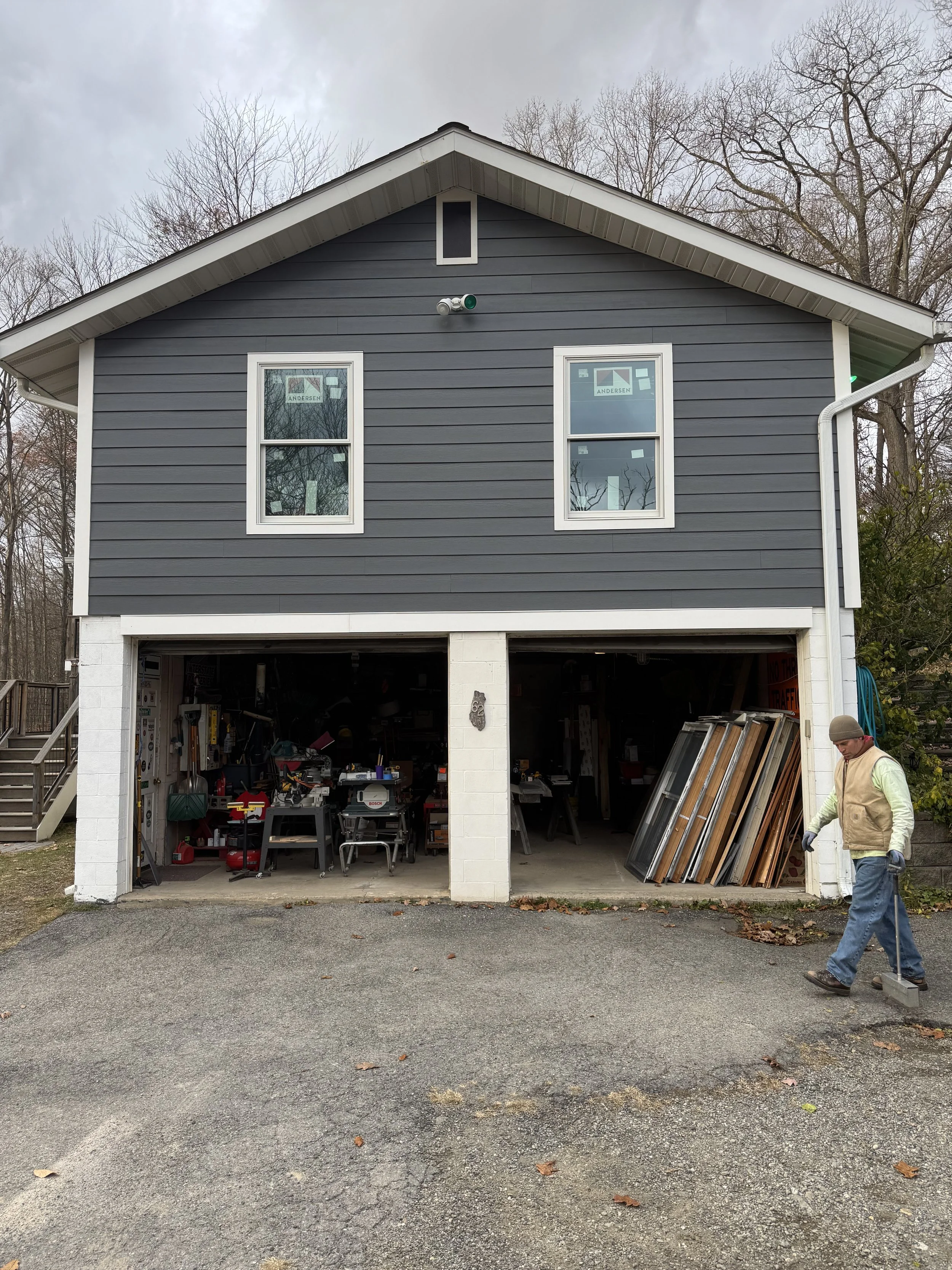 Two-car garage with the door open filled with tools and wood, a man in a vest and beanie walking outside. Bare trees and overcast sky in the background.