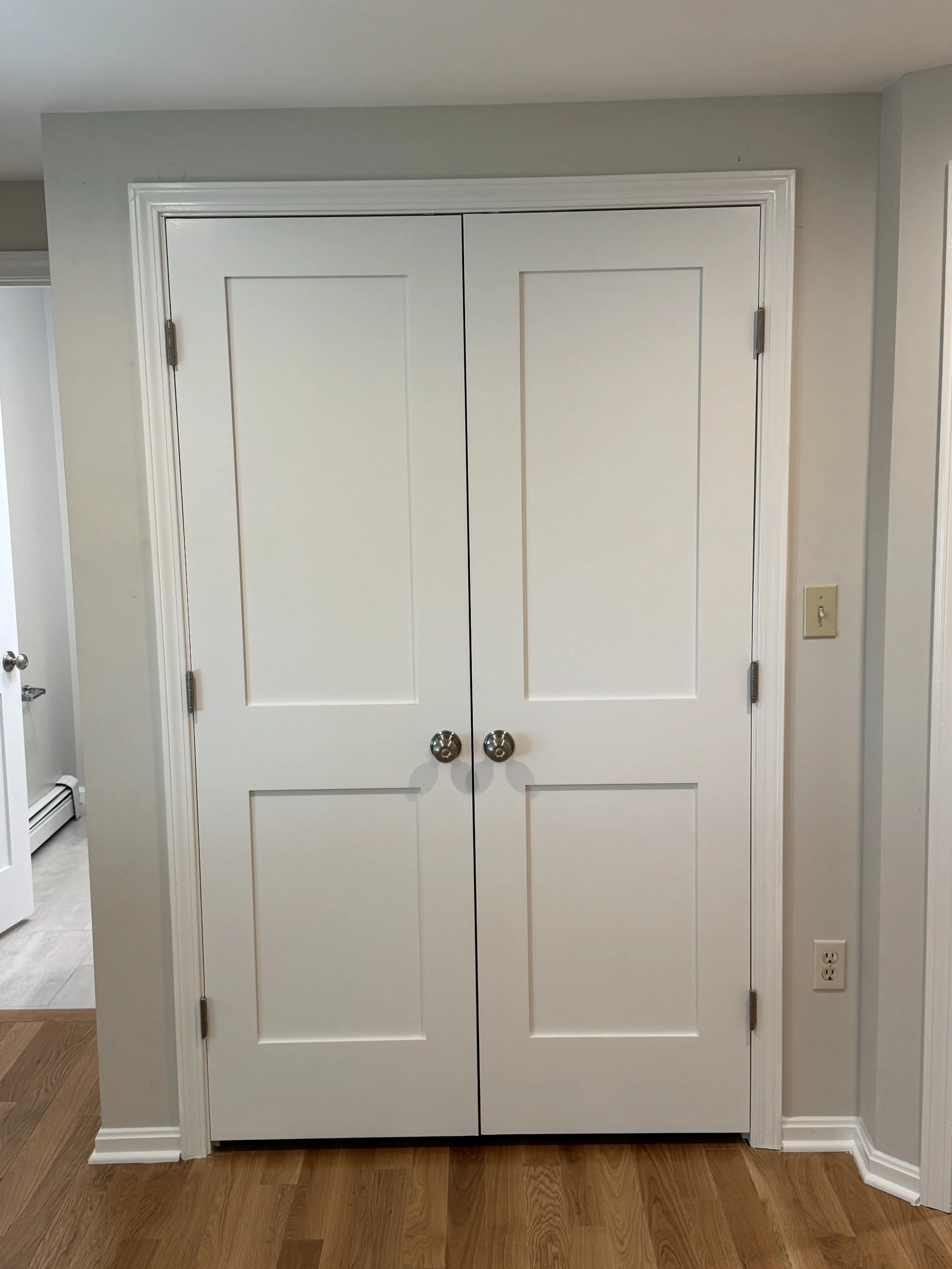 White double door closet with round silver knobs, against a wall painted light gray, with a baseboard heater and electrical outlets nearby, on a wooden floor.
