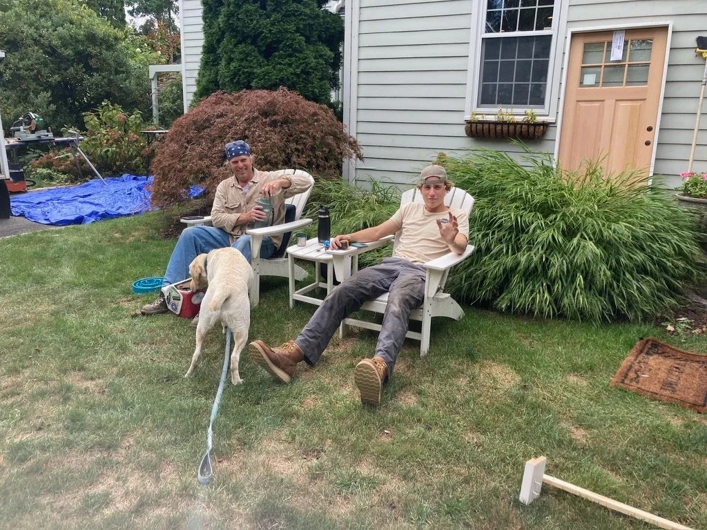 Two men sitting in white outdoor chairs in a backyard. One wears a bandana and the other a backwards cap. A yellow Labrador dog with a leash is in front, sniffing a basket. There are plants, a house with siding, and a blue tarp in the background.
