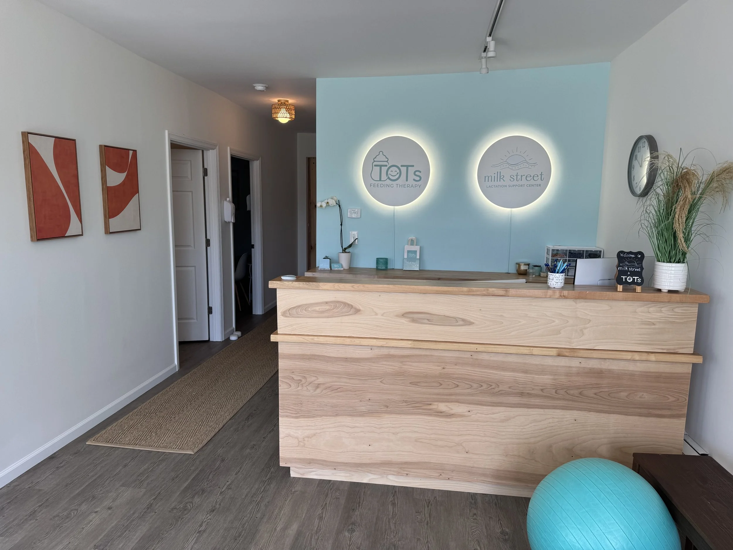 The reception area of a breastfeeding support and therapy center called TOTS feeding therapy and Milk Street Lactation Support Center, with a wooden reception desk, wall decorations, and a large clock, plus a blue exercise ball in the corner.