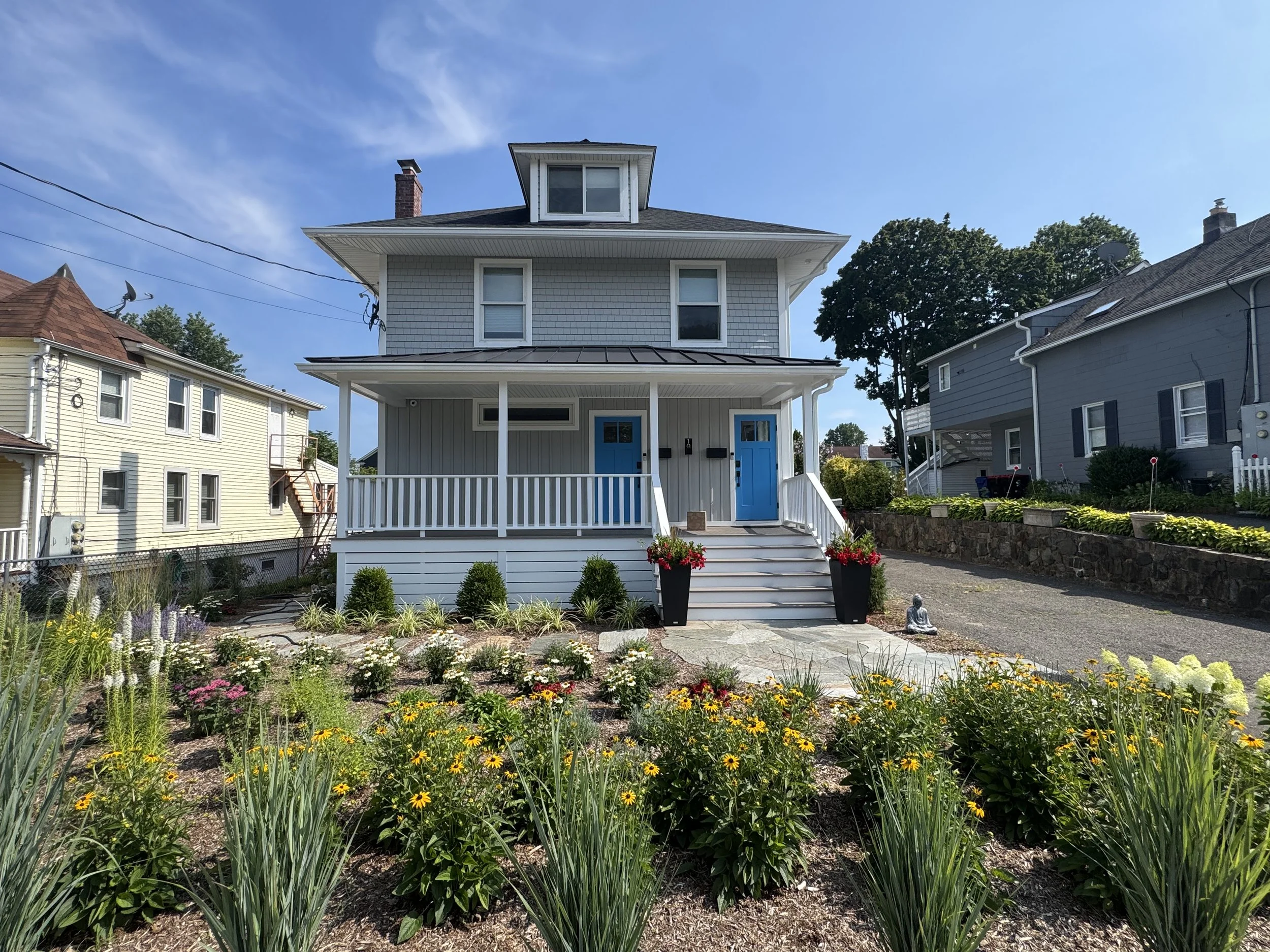A two-story house with gray siding, blue doors, and a front porch. There are flower beds with yellow and pink flowers, shrubbery, and a stone pathway leading to the house. The sky is clear with a few clouds.