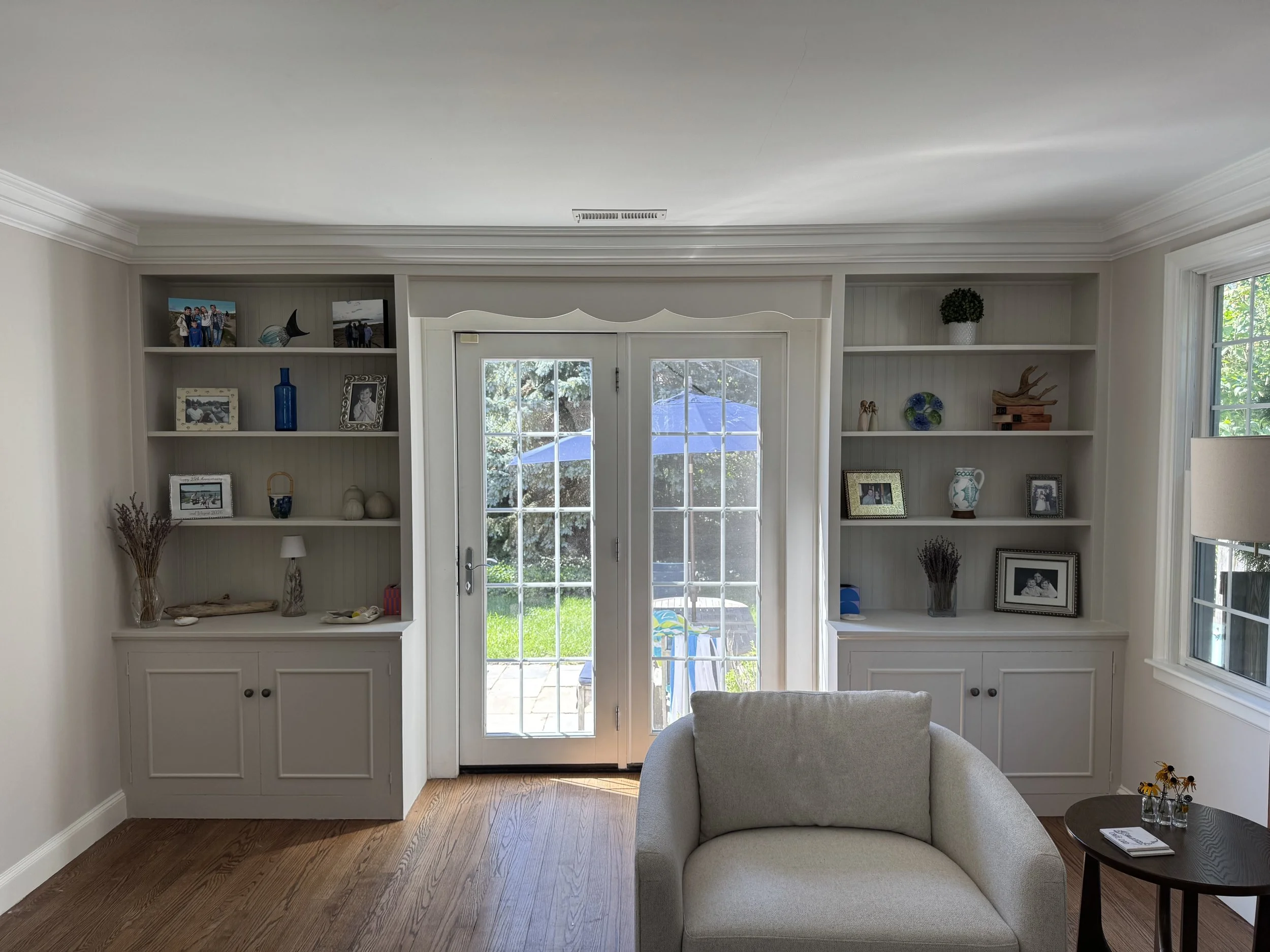 Living room with white built-in shelves, glass double doors opening to a backyard patio with an umbrella, and a beige armchair with a side table holding a glass of water and flowers.