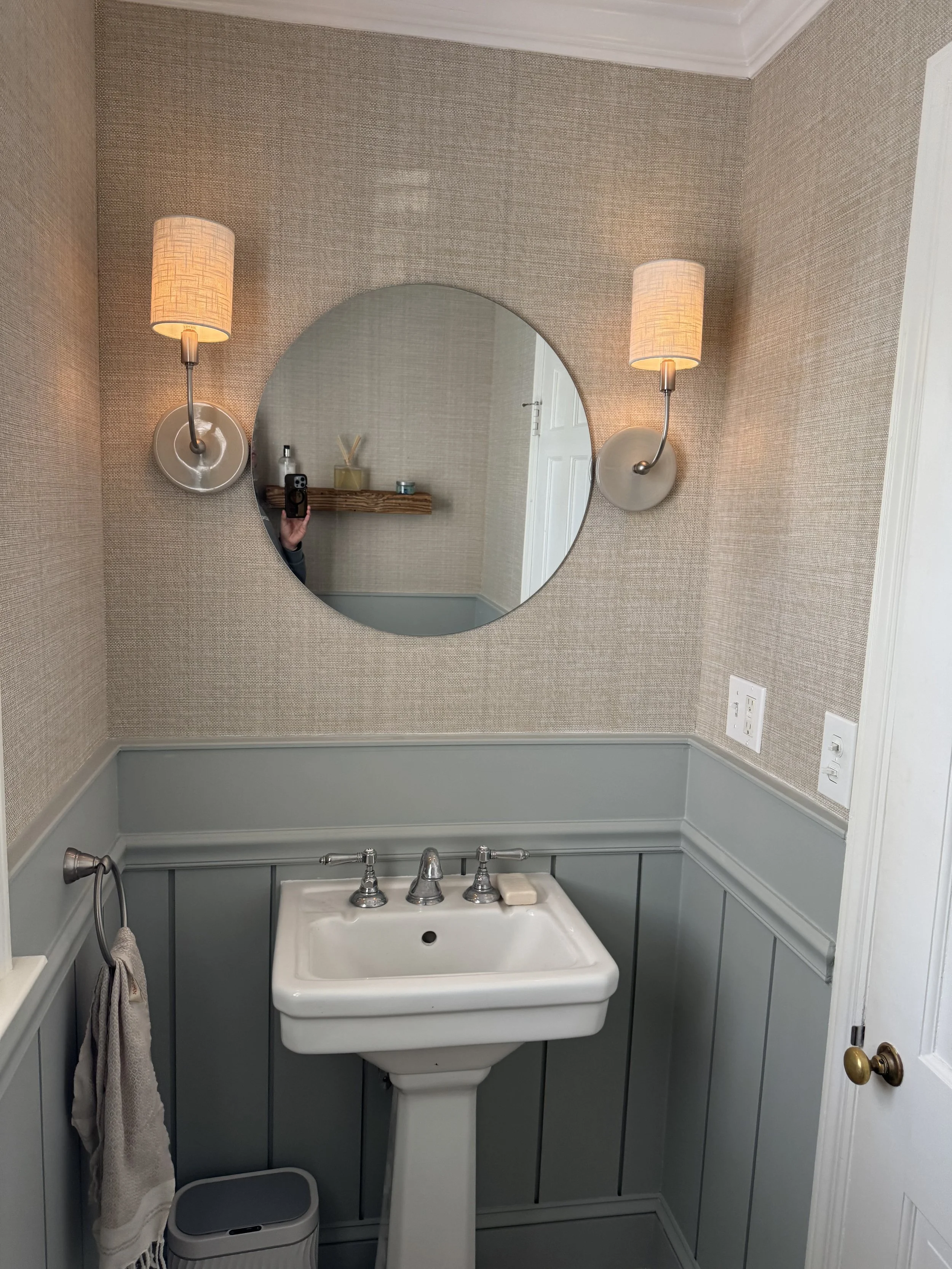 Small bathroom with a pedestal sink, oval mirror, beige textured wallpaper, two wall-mounted lamps with shade coverings, a towel hanging on a hook, and a small trash can on the floor.