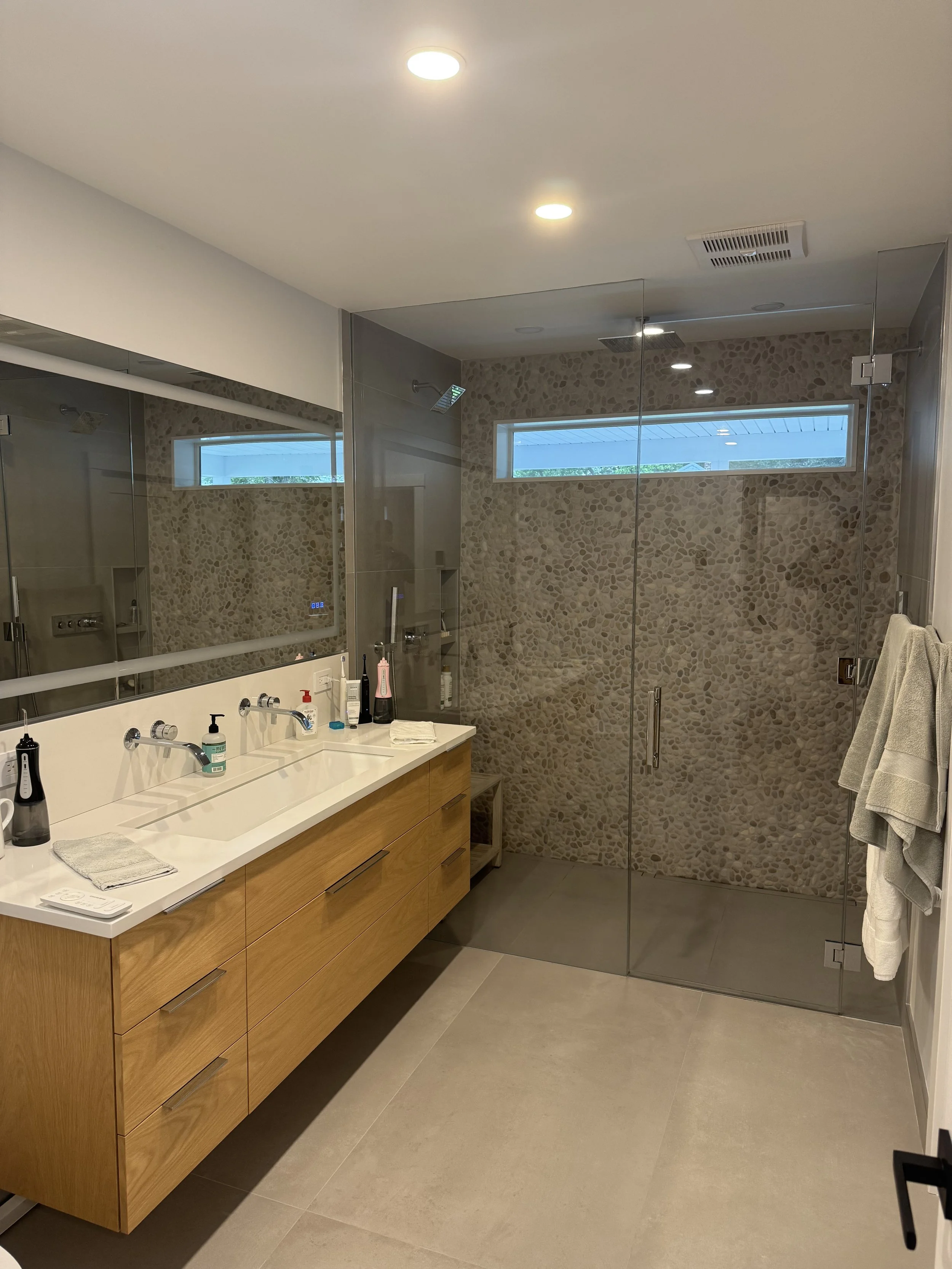 Modern bathroom with wooden vanity, large mirror, and glass-enclosed shower with pebble wall and horizontal window.