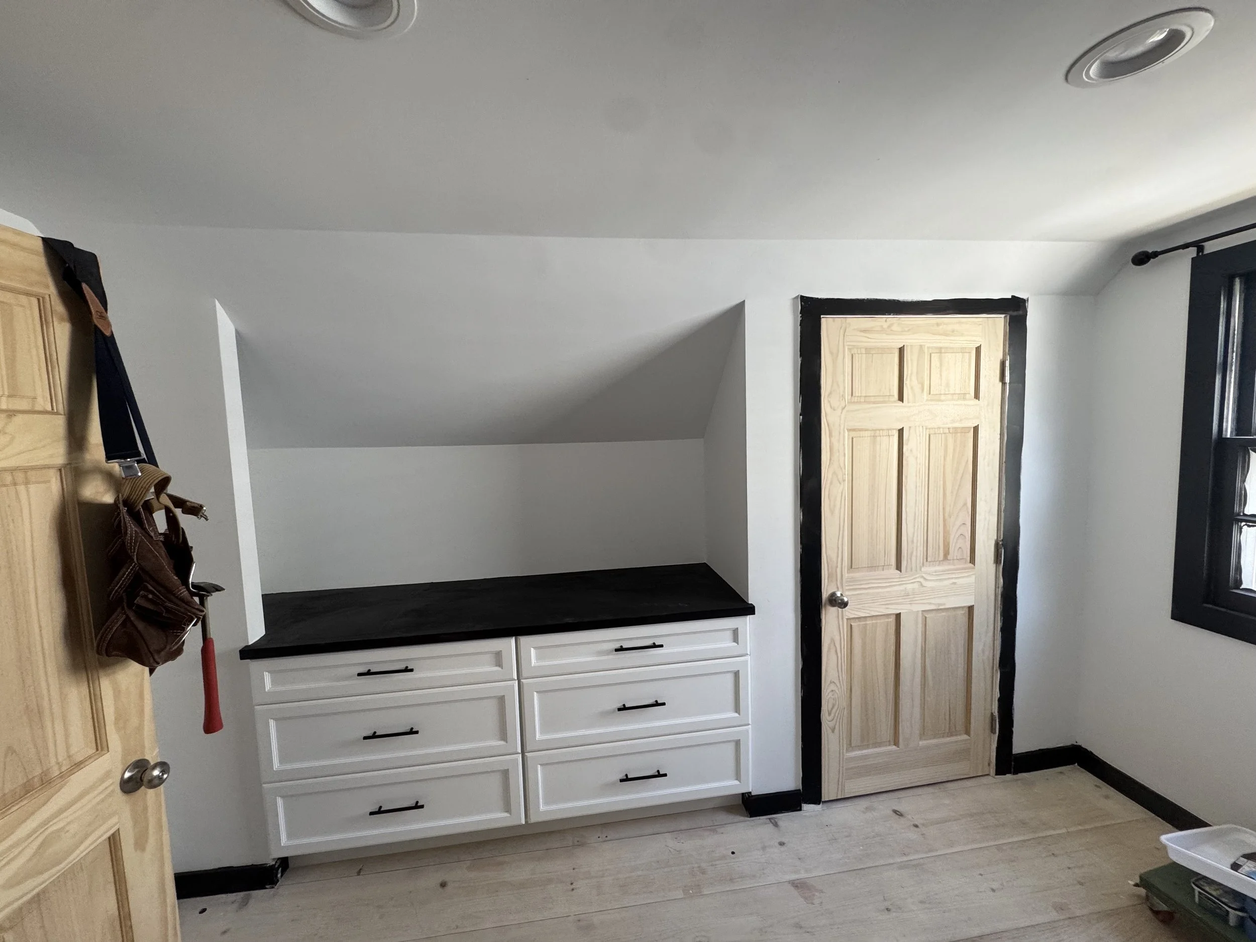 Interior view of a room with white cabinets, black countertop, wooden door, window with black frame, and a sloped ceiling.