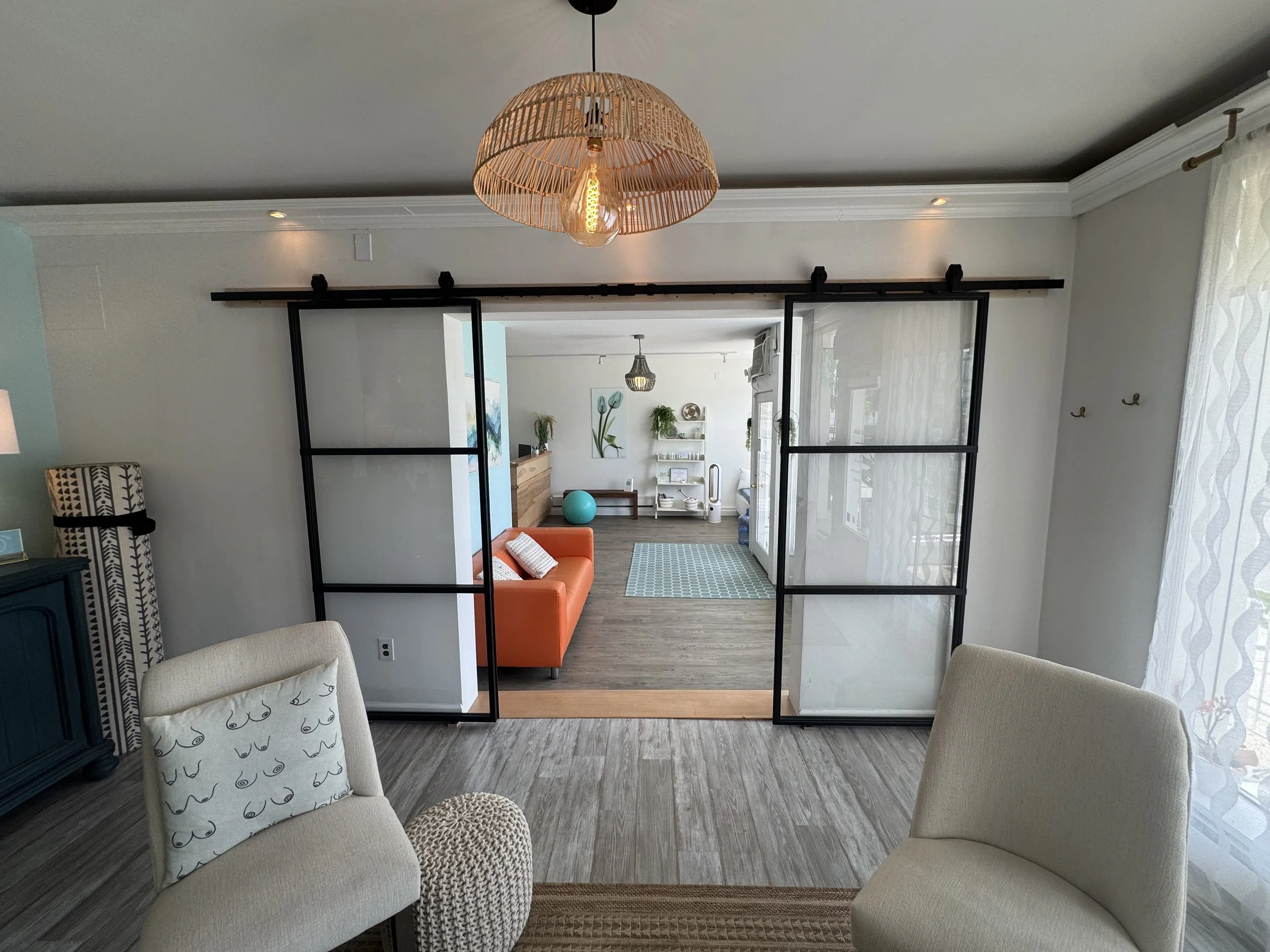 A living room with sliding barn doors, an orange couch, patterned pillows, a small patterned ottoman, and wall art, seen through open doors with a view of another room.