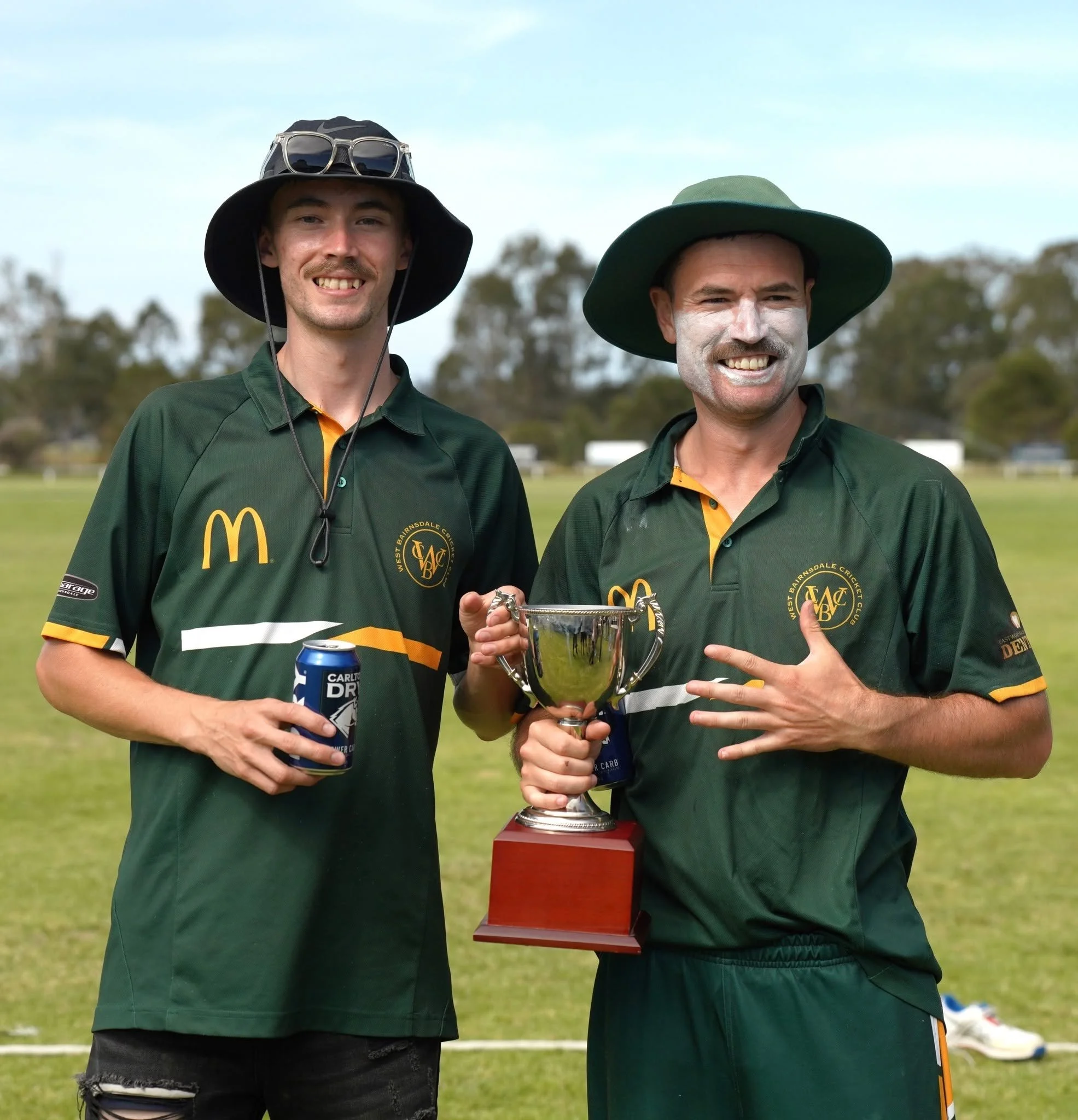 Two men standing outdoors on a sports field, smiling; the man on the left wears a leaf-patterned hat with sunglasses on top, and the man on the right wears a broad green hat with white face paint, holding a silver trophy. Both wear green sports jerse