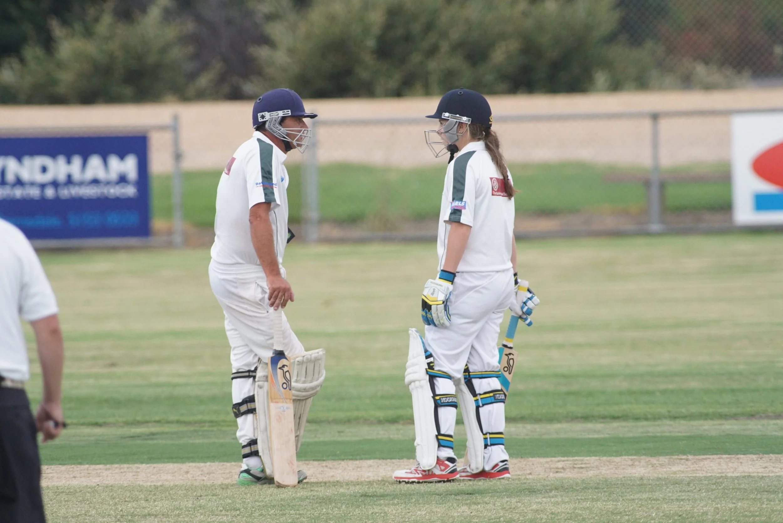 Two cricket players, a man and a woman, standing on the field in cricket gear, engaged in conversation during a game.