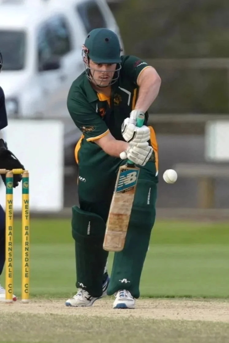 Cricket batter in green uniform, wearing a helmet, pads, and gloves, hitting the ball with a cricket bat on a cricket field.