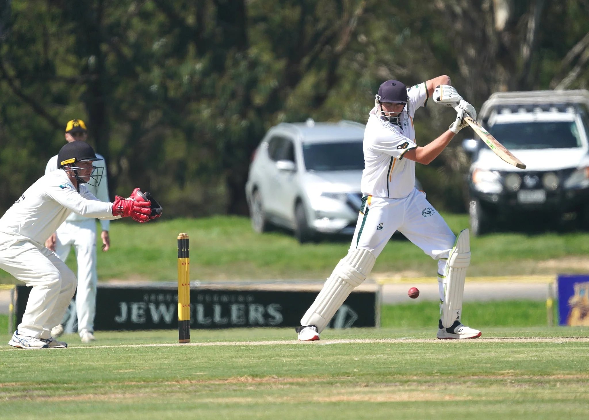 Cricket players in action on the field, with one batsman hitting the ball while two fielders and a wicketkeeper are positioned nearby.