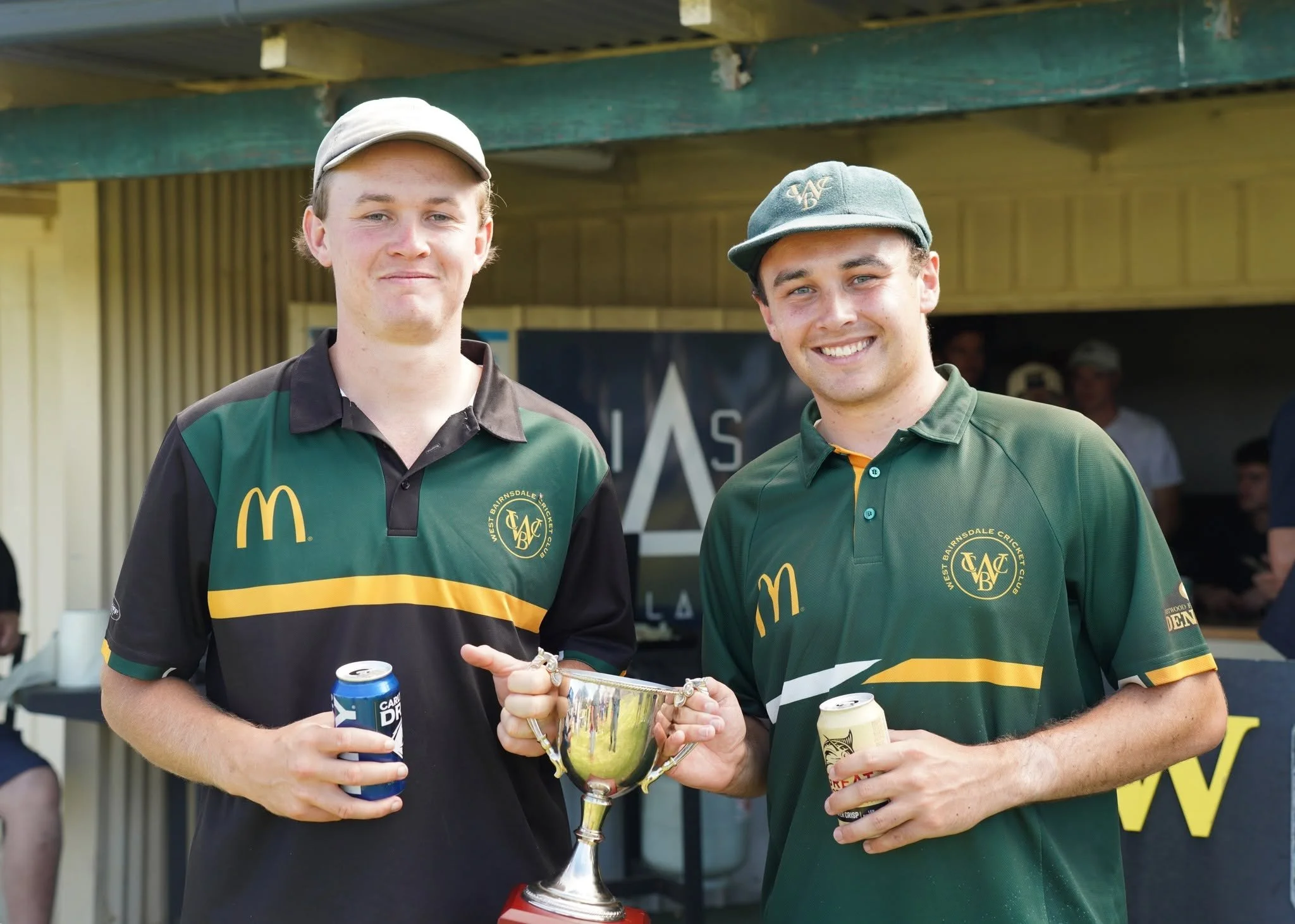 Two young men in sports uniforms smiling and holding a trophy and drinks, standing outdoors at a sporting event.