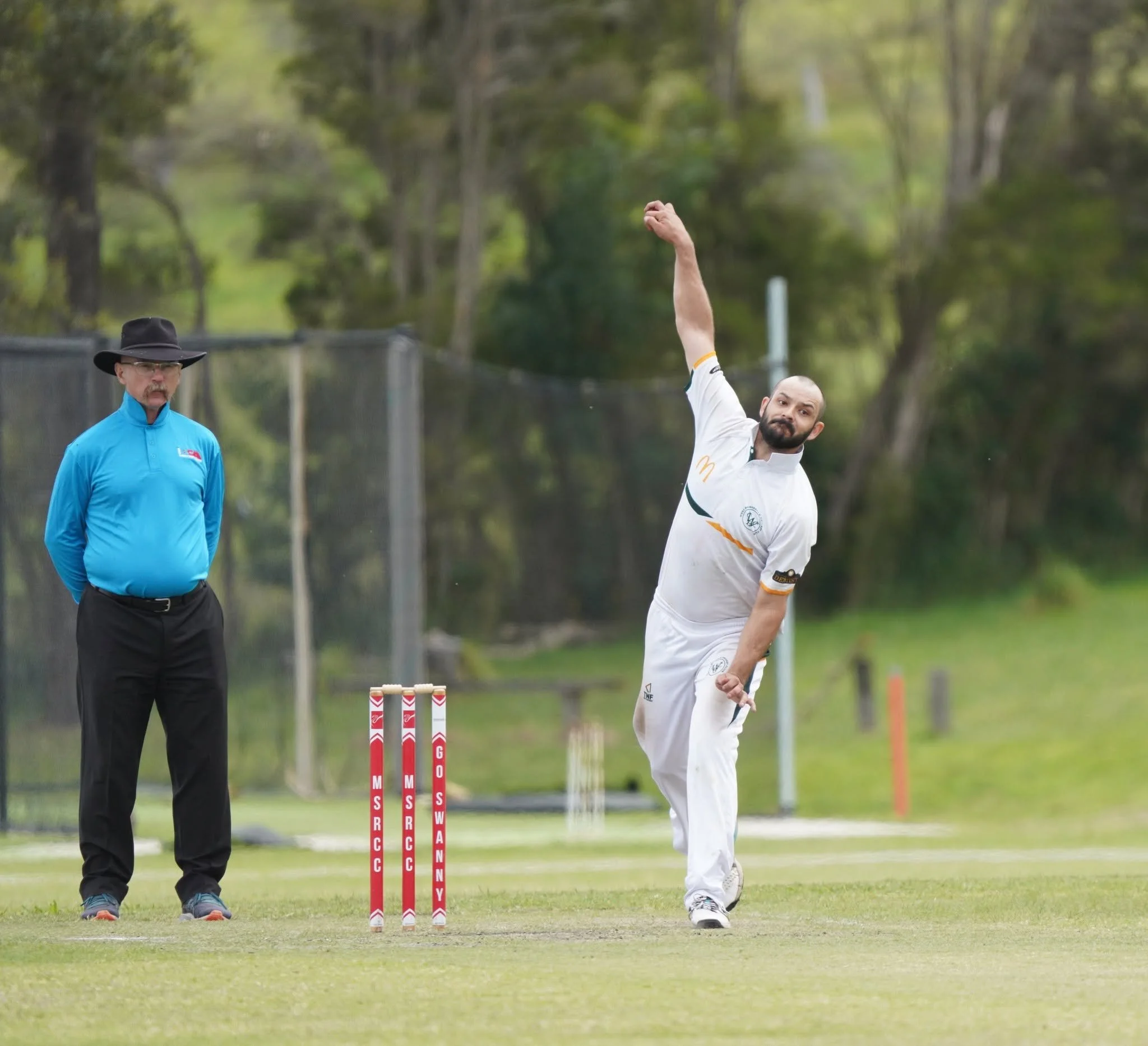 Cricket player in white uniform celebrating with raised fist on field during game, umpire in blue shirt watching, wickets behind.