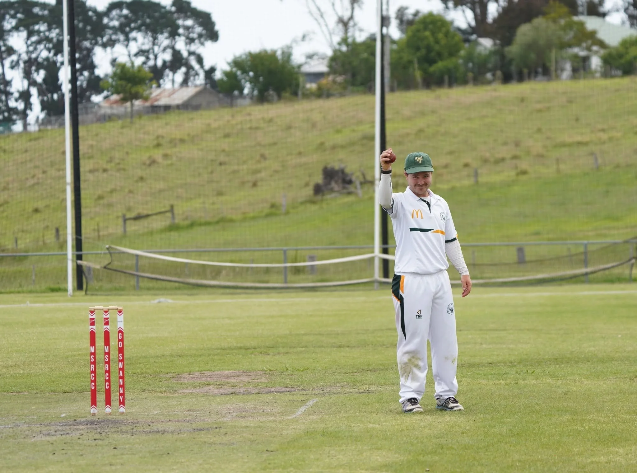 A man wearing a green cap and white sports uniform with logos and text stands on a cricket field, smiling and waving his right hand. In the background, four red cricket stumps are arranged with the stump pegs labeled 'MSRC C', 'MSRC C', and 'W A N Y'
