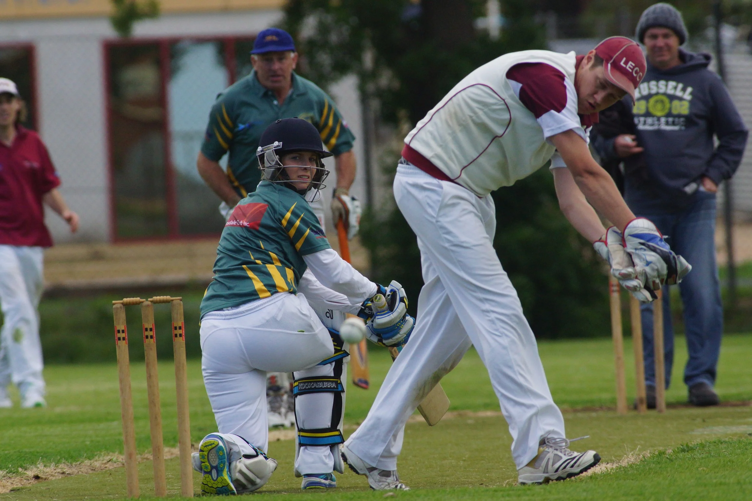 A cricket match scene with a young player bowing and in wicket-keeping position, others observing, on a grassy field.
