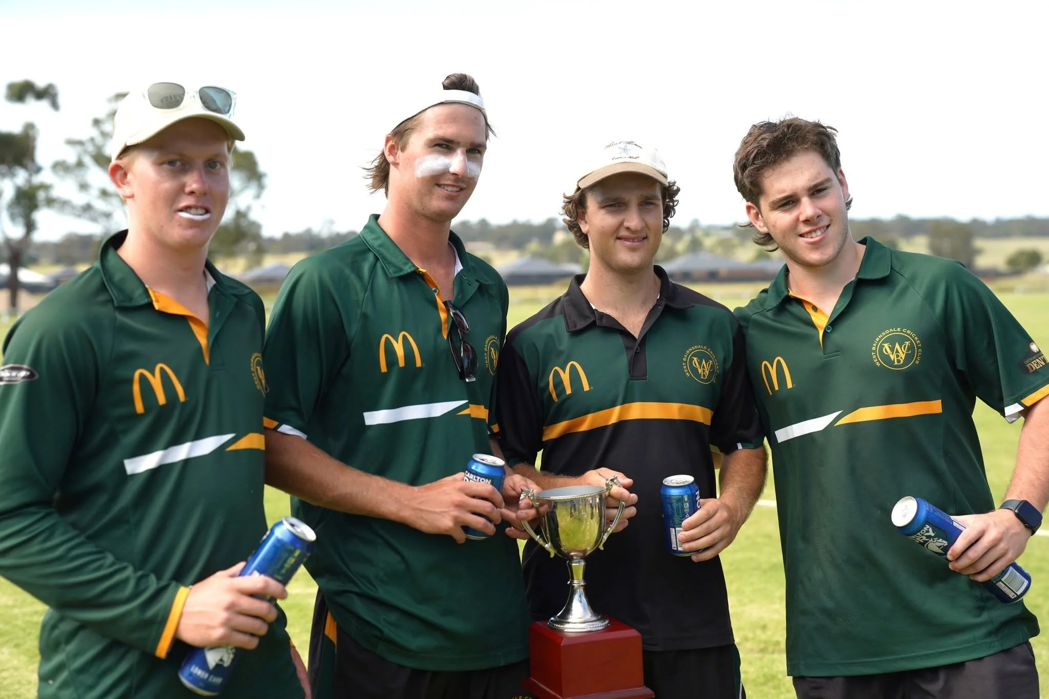 Four young men in green and black sports jerseys standing outdoors on a grassy field, holding beverages, with a trophy in the center, smiling at the camera.