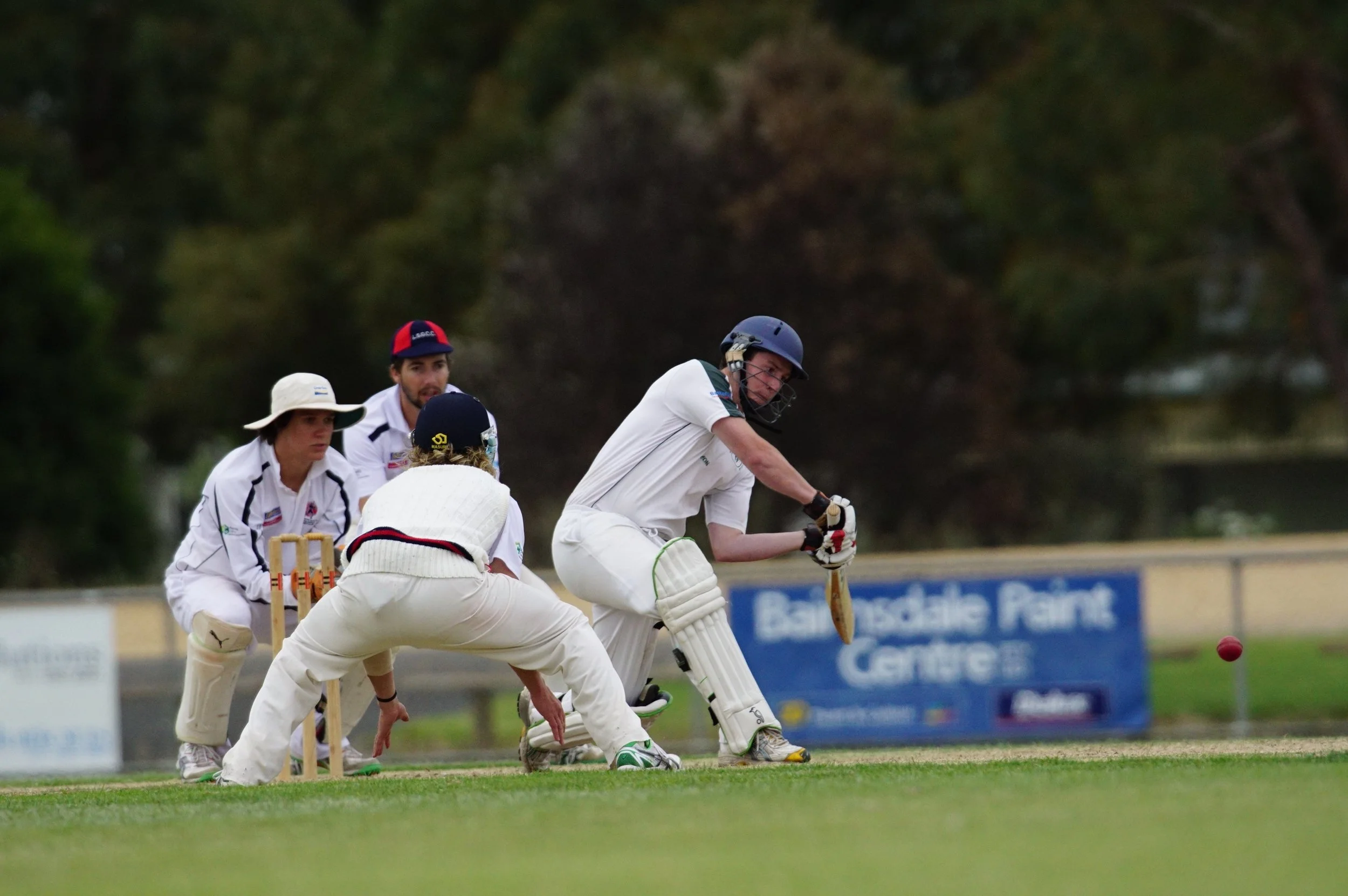 Cricket players in white uniforms playing on a field with a wicket and a red ball in play. One player is bending down to hit the ball while others are observing.