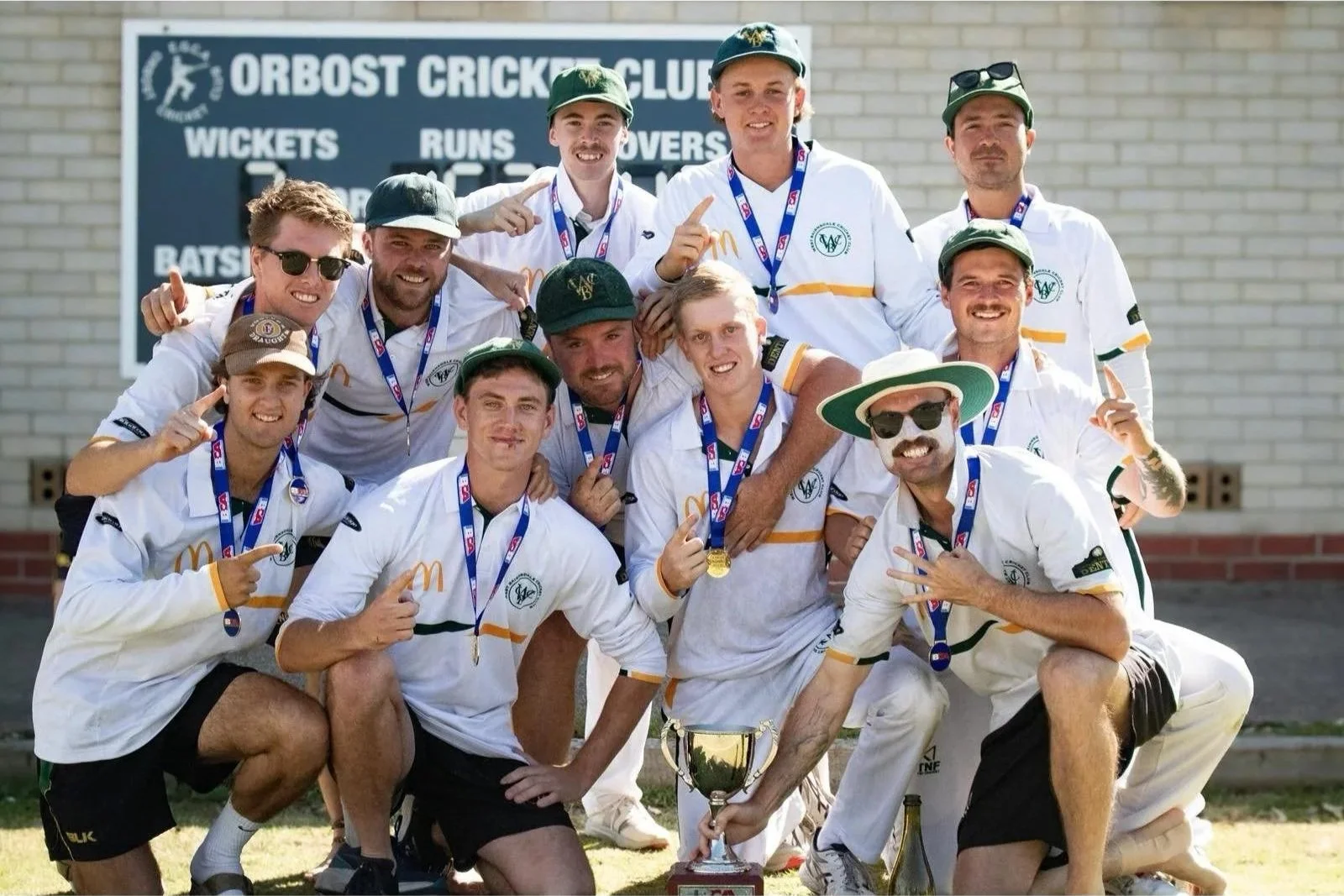 Cricket team celebrating with medals and trophy at Orbst Cricket Club, standing in front of a signboard with team statistics.