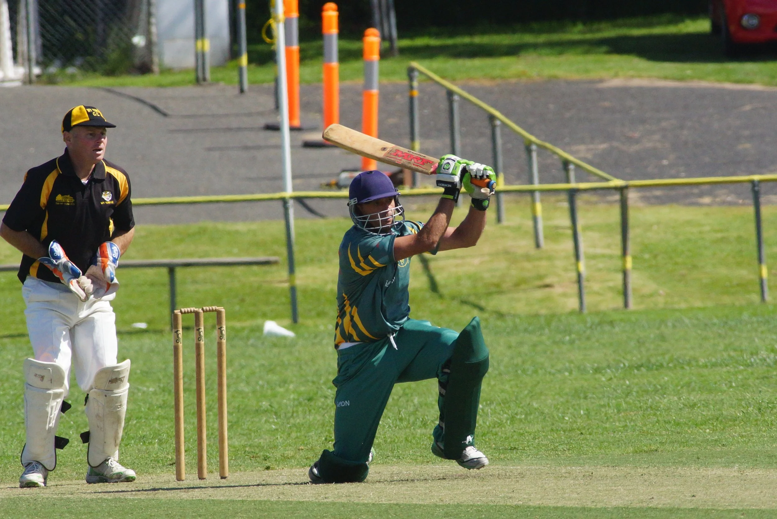 Cricket player in a teal uniform and helmet is swinging a bat during a game while a wicket keeper wearing protective gear stands behind him and another player watches nearby on a grassy field with orange and yellow poles and fencing in the background