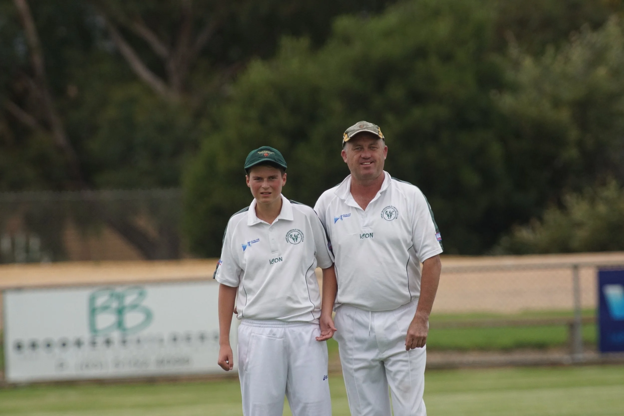 Two men in white cricket uniforms walking outdoors on a grassy field, with trees in the background.