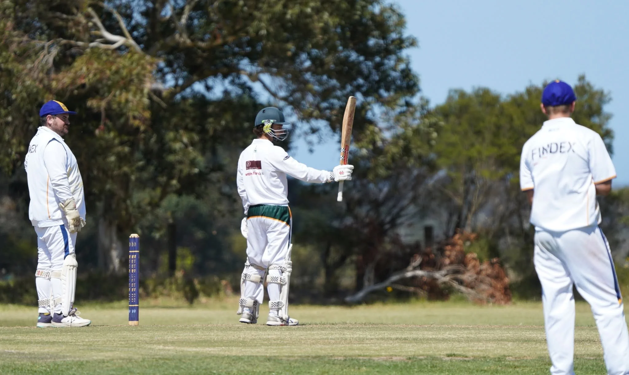 Cricket players on the field with one holding a cricket bat and wearing protective gear, standing near wickets, with trees in the background.