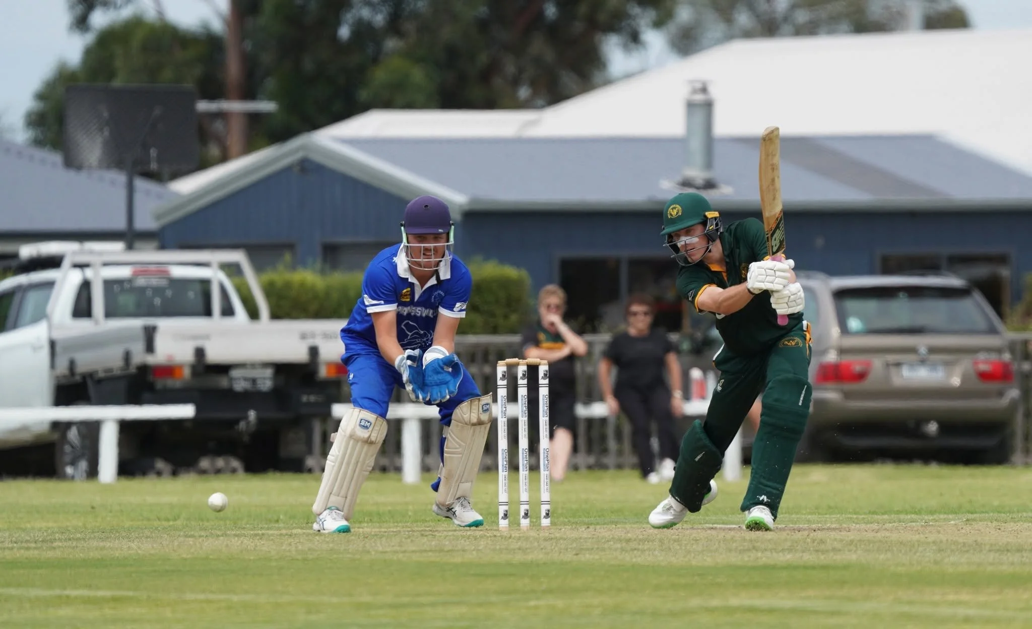 A cricket bowler in a green uniform and helmet is mid-action, delivering the ball, while a wicketkeeper in a blue uniform and helmet crouches behind the stumps on the cricket field. Spectators and parked cars are visible in the background.