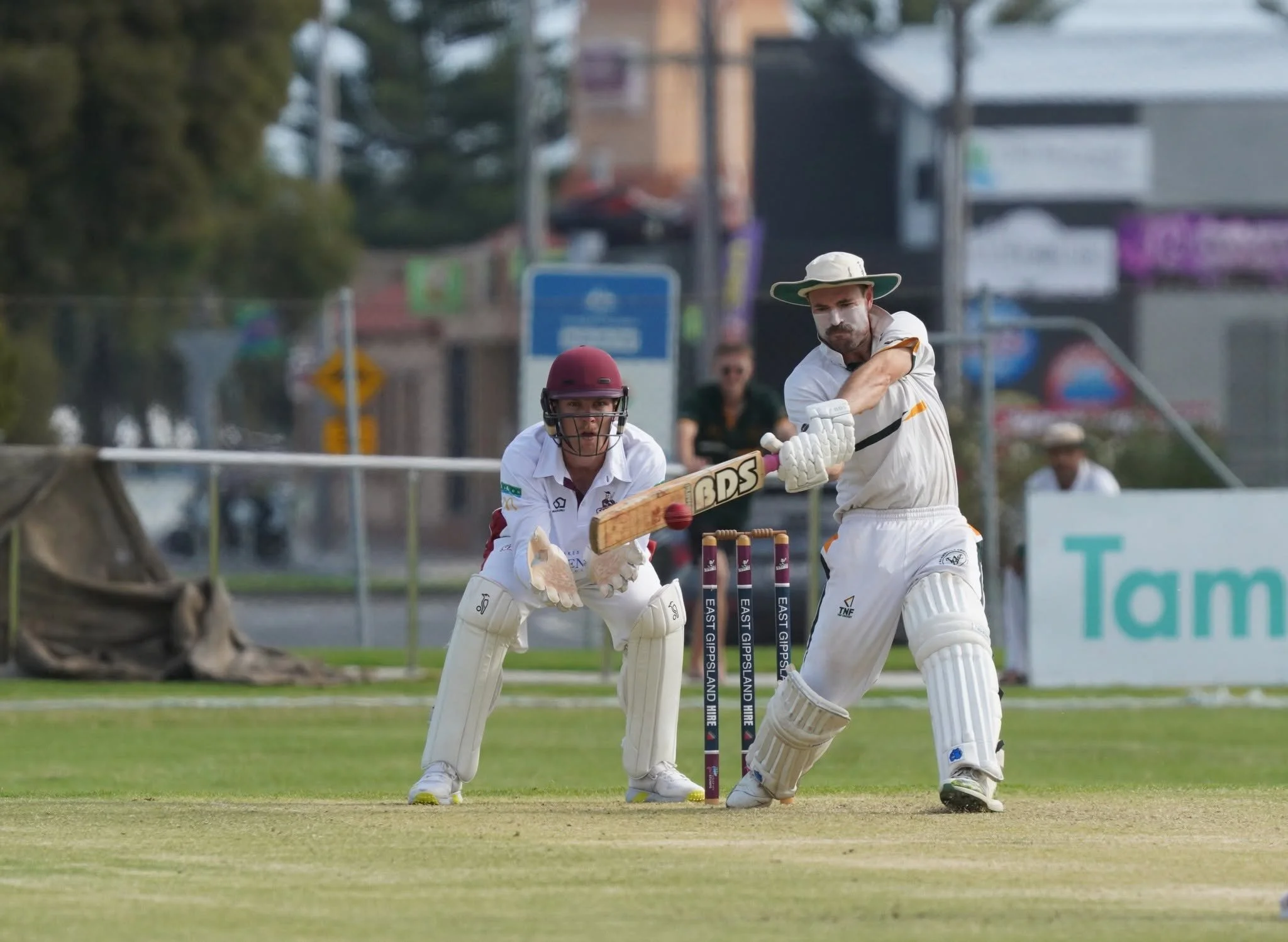 Two cricket players in white uniforms and protective gear, including helmets, playing on a cricket field during daytime. One player is batting, while the other is fielding behind him.