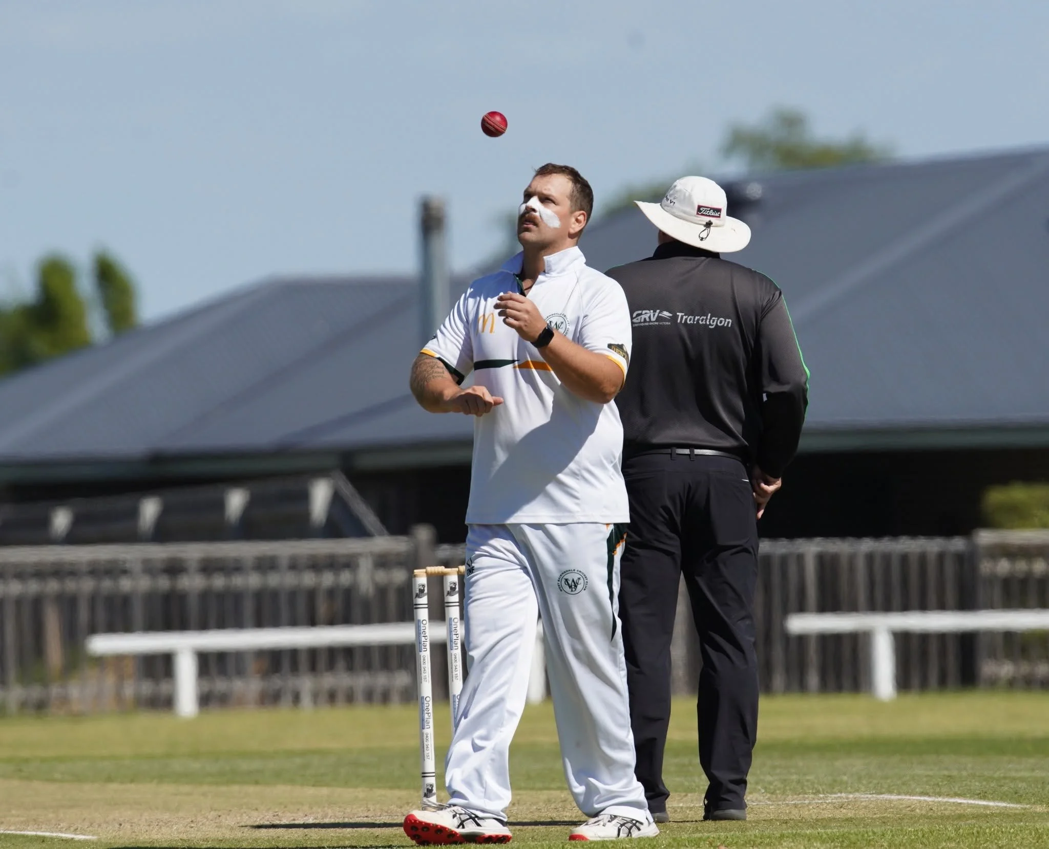 A man in cricket attire with face paint on his nose, next to a man wearing a hat, standing on a cricket field, with a red cricket ball in mid-air above them.