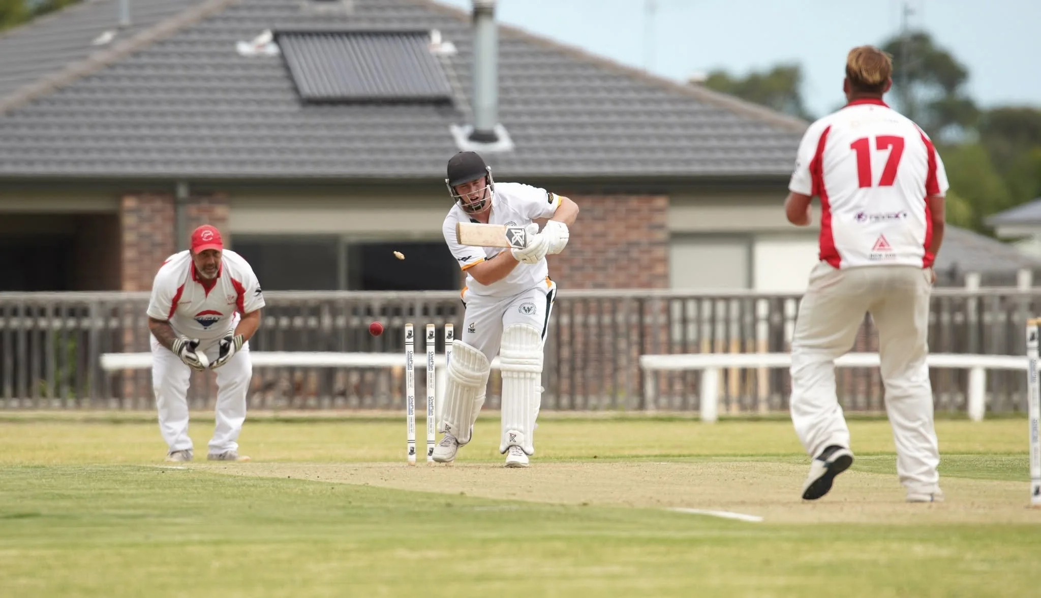 Cricket players during a match; one player is batting while two other players are on field, with a house and fence in the background.