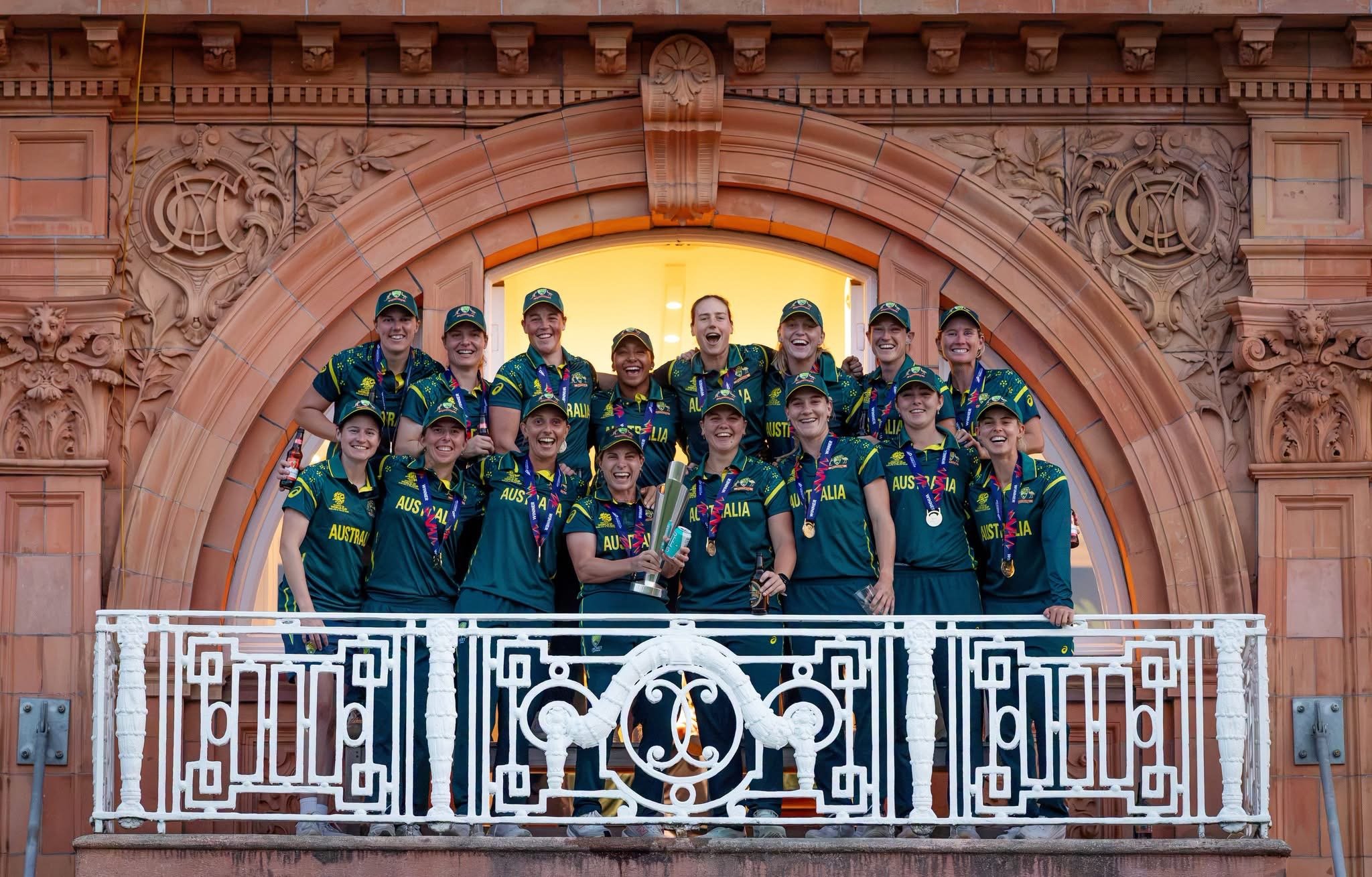 A group of women dressed in green and yellow sports uniforms standing on a balcony with ornate brickwork. They are celebrating, smiling, and some are holding medals and a trophy. The scene appears to be a celebration or victory moment.