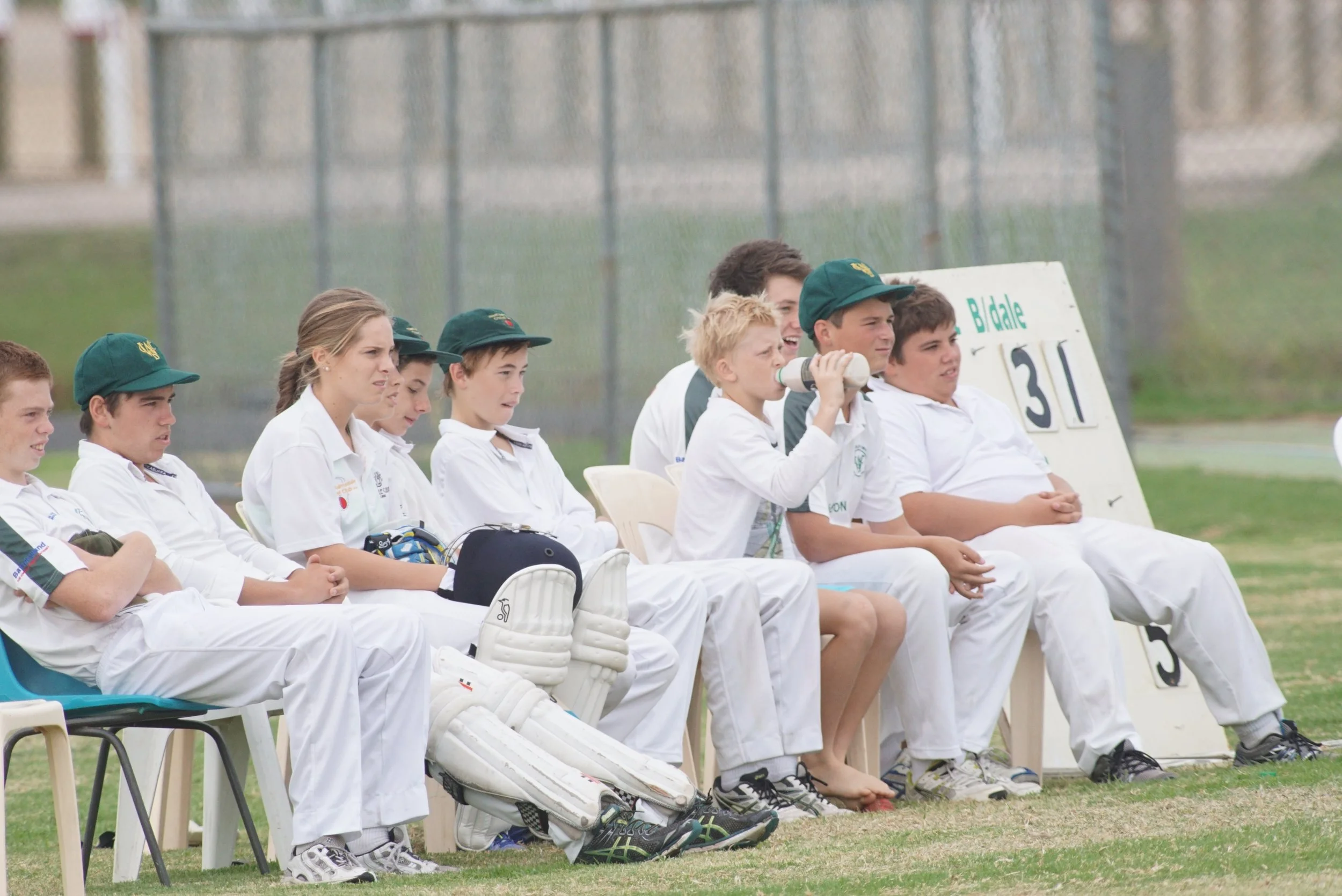 A group of young cricket players sitting on a bench during a game, some wearing white cricket uniforms and green caps, with a scoreboard and chain-link fence in the background.