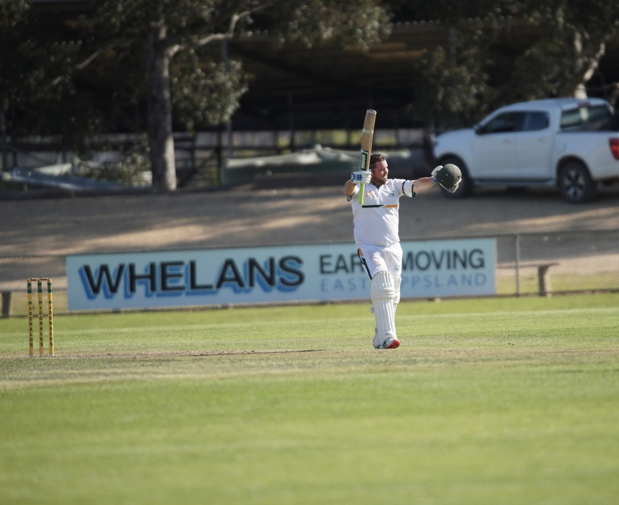 A cricket player celebrating on the field after a successful shot, holding a helmet in one hand and a cricket bat raised in the other, during daytime, with boundary markers and advertising signs visible in the background.