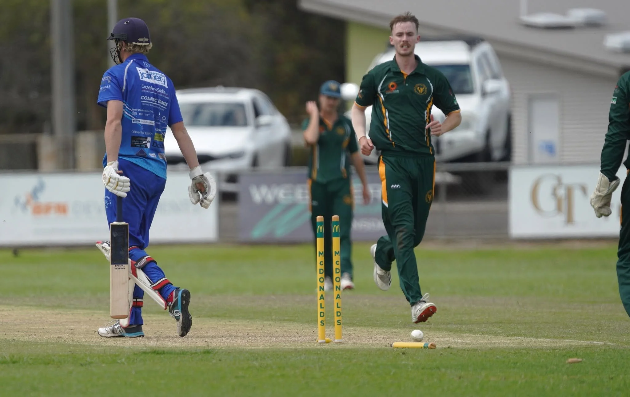Cricket players celebrating on the field, with a focus on a player in green uniform running past a stumps, and a wicket-keeper in blue uniform standing nearby, during a match.