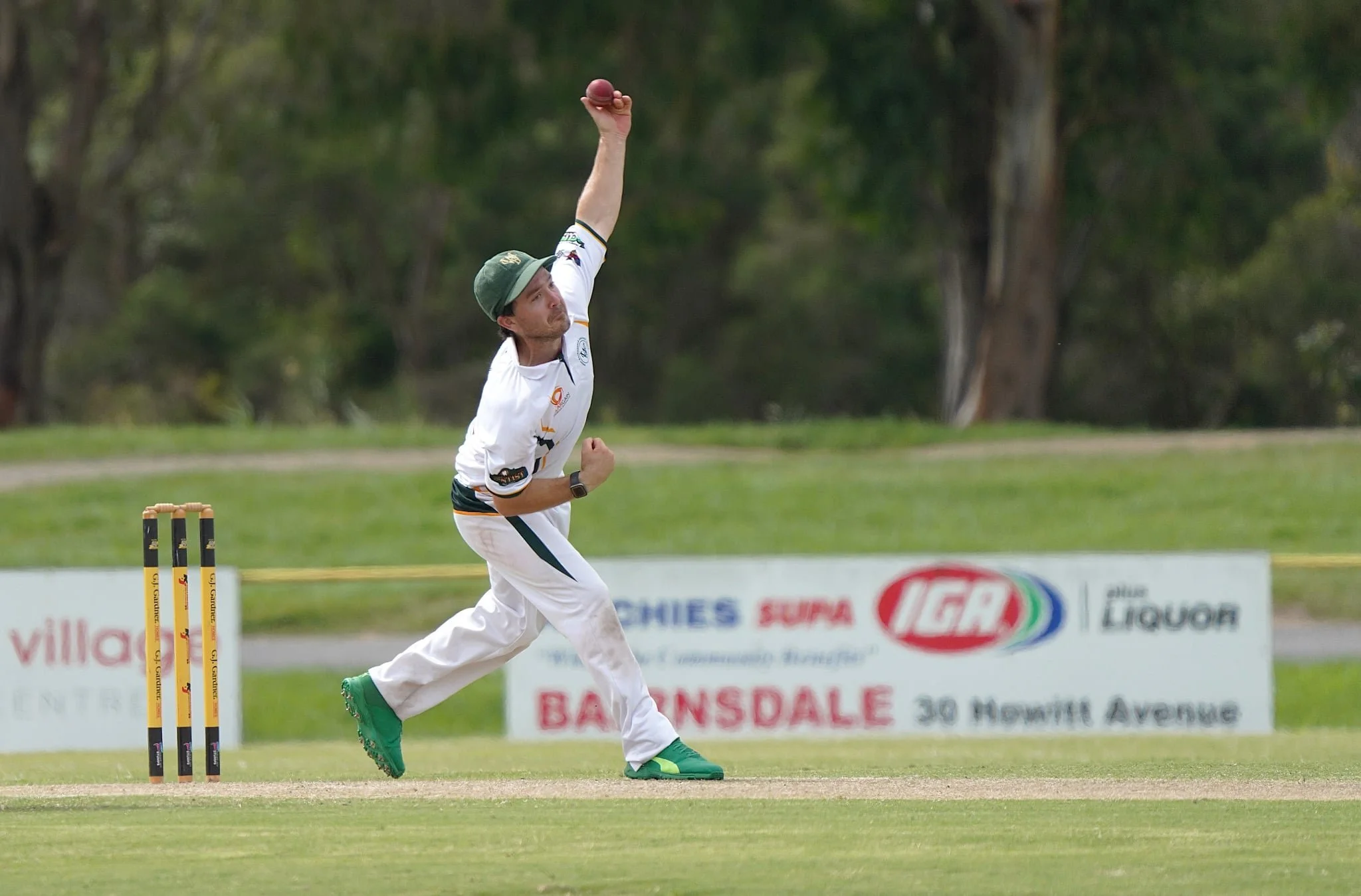 A cricket player wearing a white uniform with green shoes and a green hat celebrating a successful catch on the cricket field, with trees and advertising boards in the background.