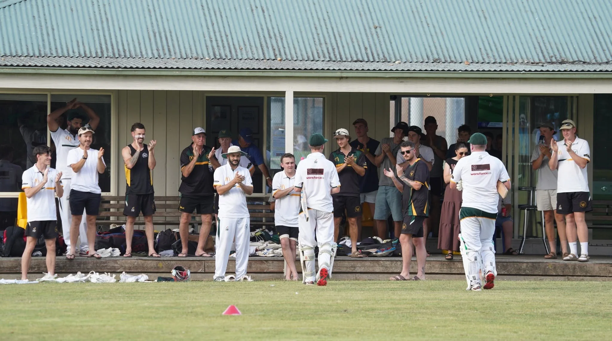Cricket team and spectators on a pavilion during a match, some players are clapping and standing, with cricket equipment on the ground.