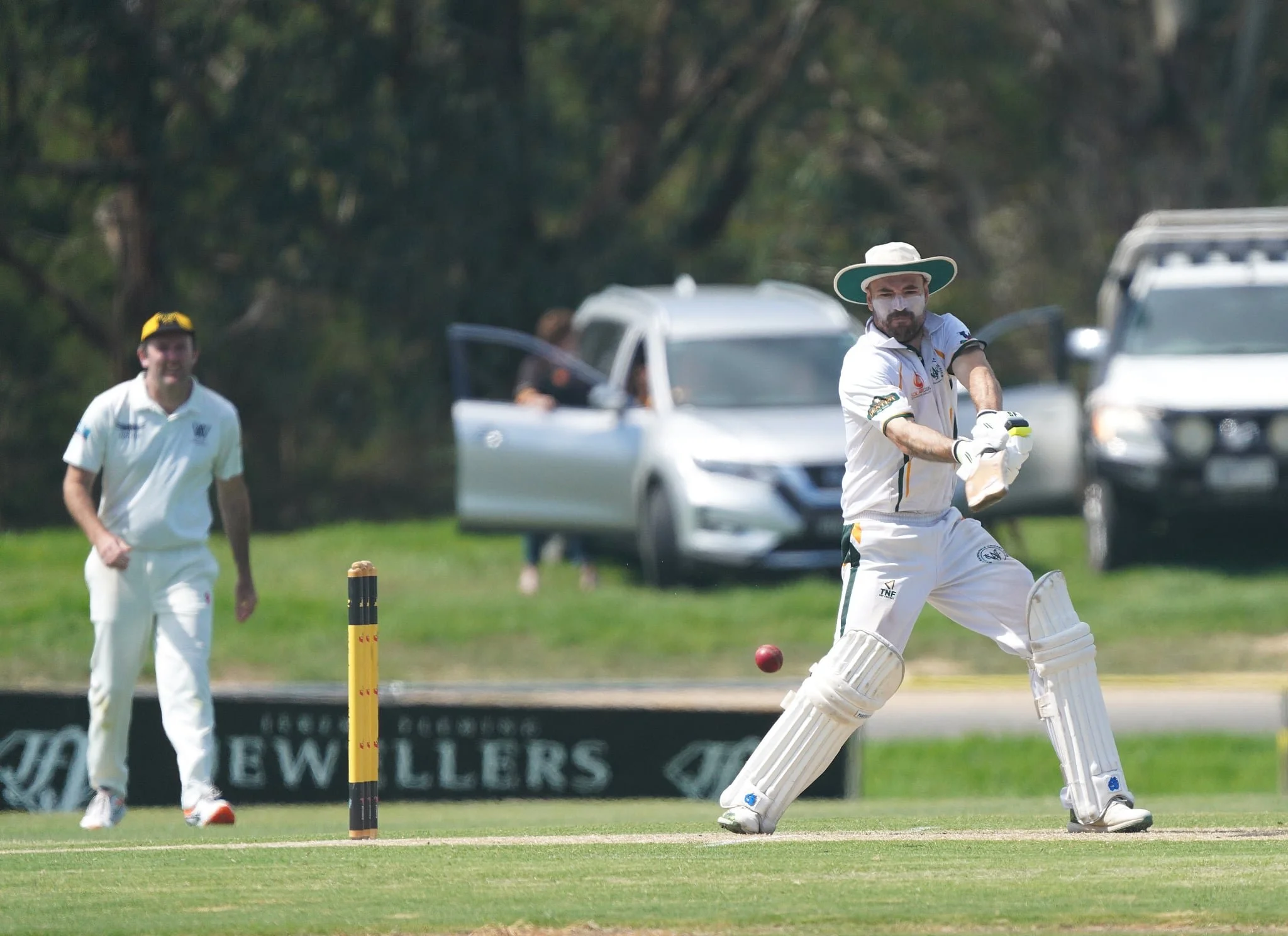 A cricket player in white uniform and large sun hat is about to hit the red cricket ball with a bat. Two other players are visible in the background, one on the field and the other near the parked cars, with greenery and trees behind them.