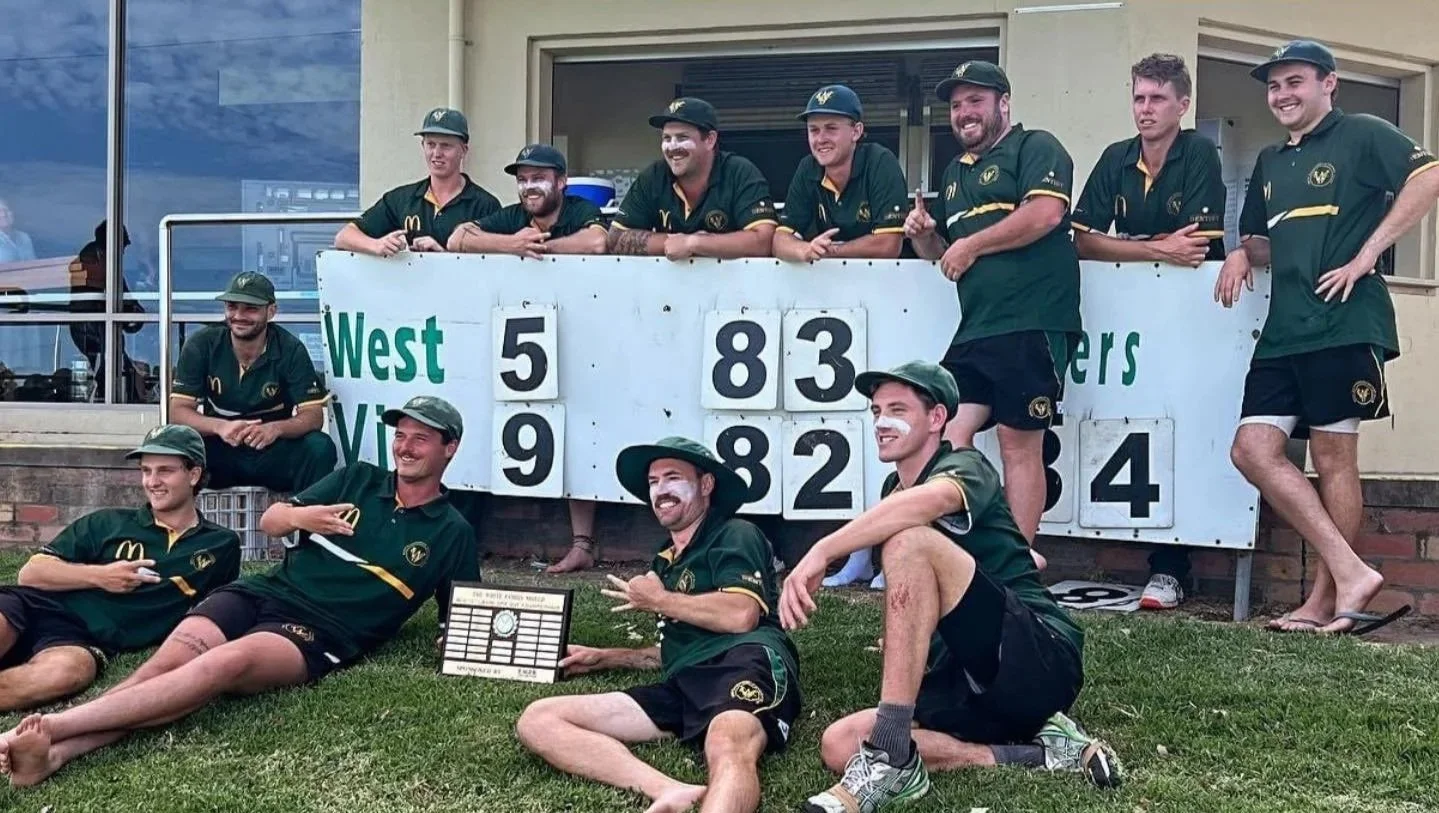 A group of men in green sports uniforms with face paint, some lying on the grass and some standing, posing with a scoreboard showing run scores, celebrating after a sports game.