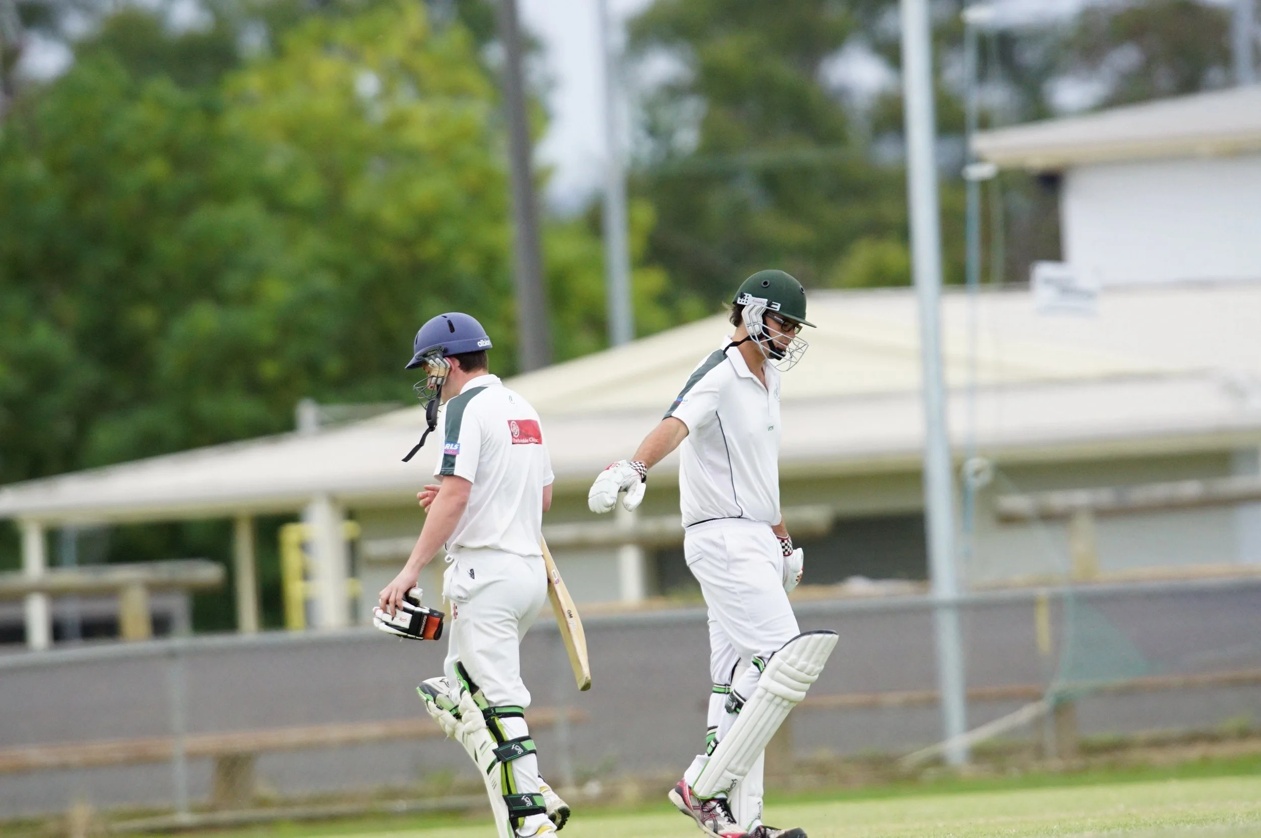 Two cricket players in white uniforms walking on a cricket field, wearing helmets, with a blurred background of trees and a fence.
