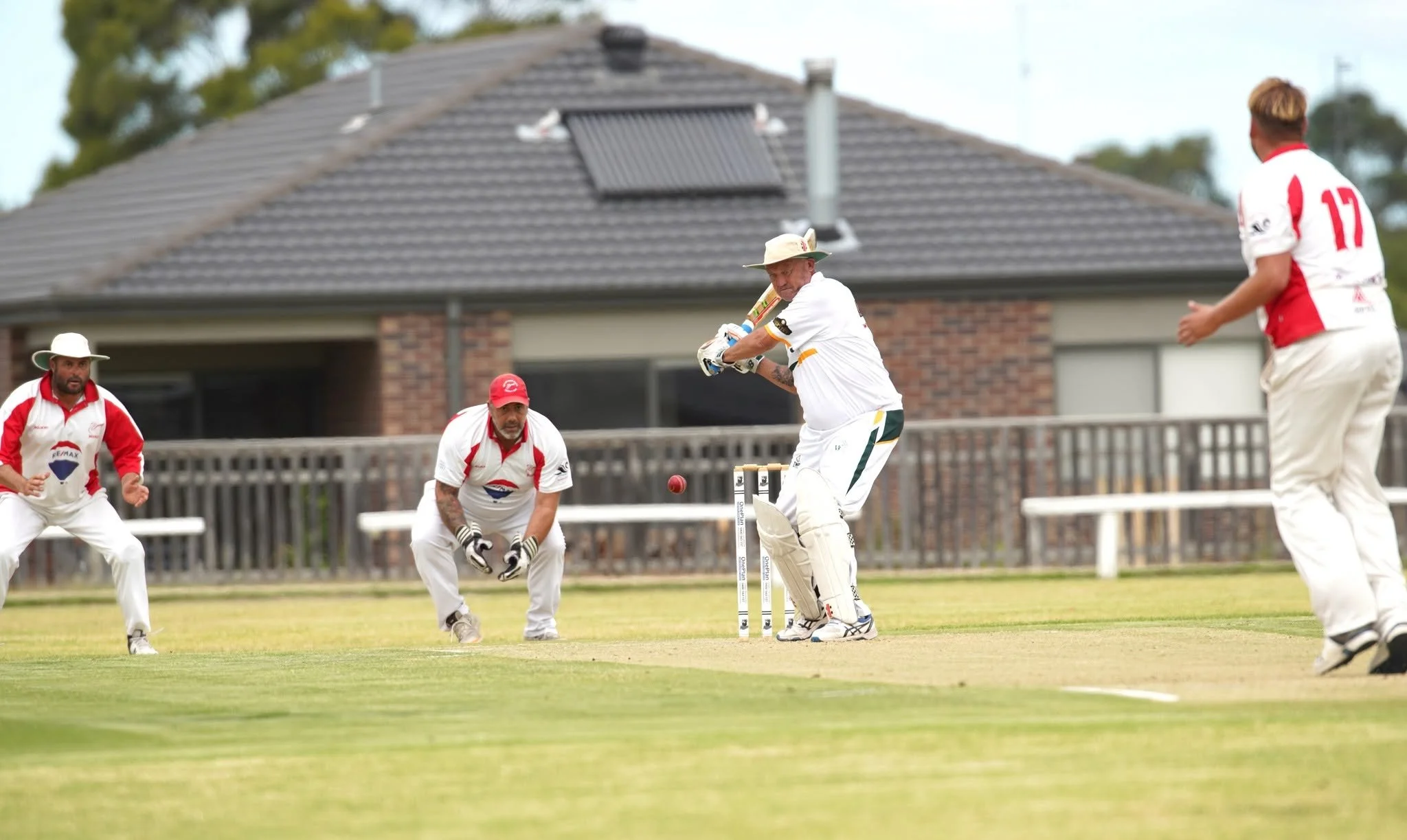 Cricket players in white and red uniforms on the field, focusing on a batsman preparing to hit the ball, while other players are in fielding positions behind him, with a house and trees in the background.