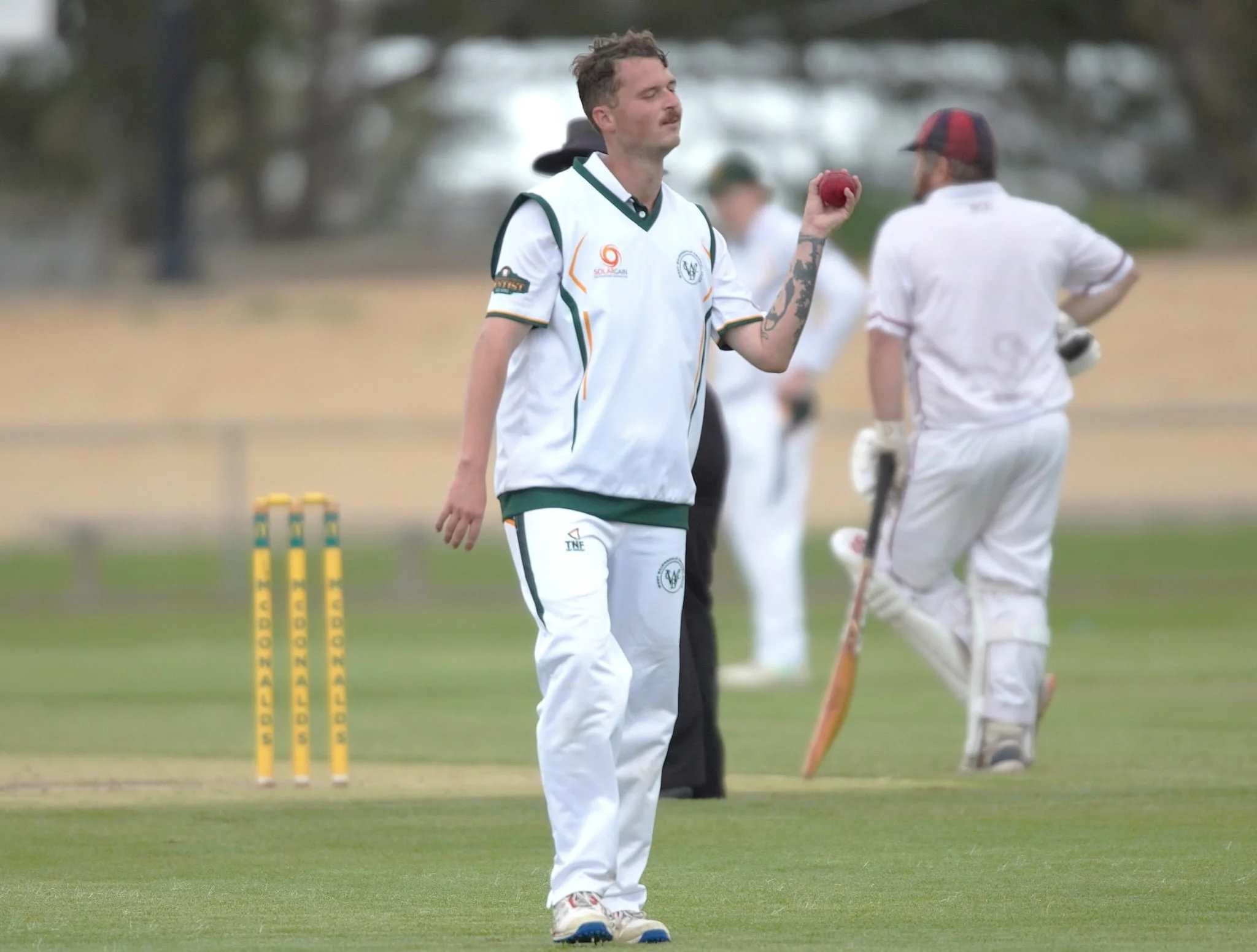 A cricket player in a white uniform holding a red cricket ball on a cricket field. Other players and the stumps are visible in the background.