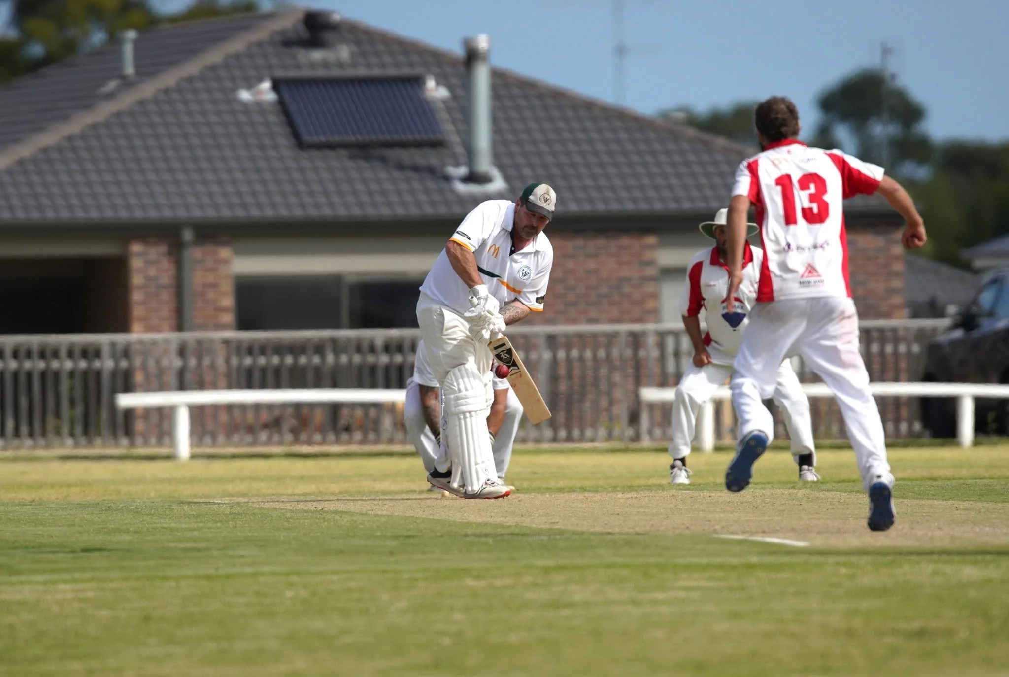 Cricket players on the field during a game, with one player preparing to strike the ball, while others watch, and a house with a solar panel on the roof in the background.