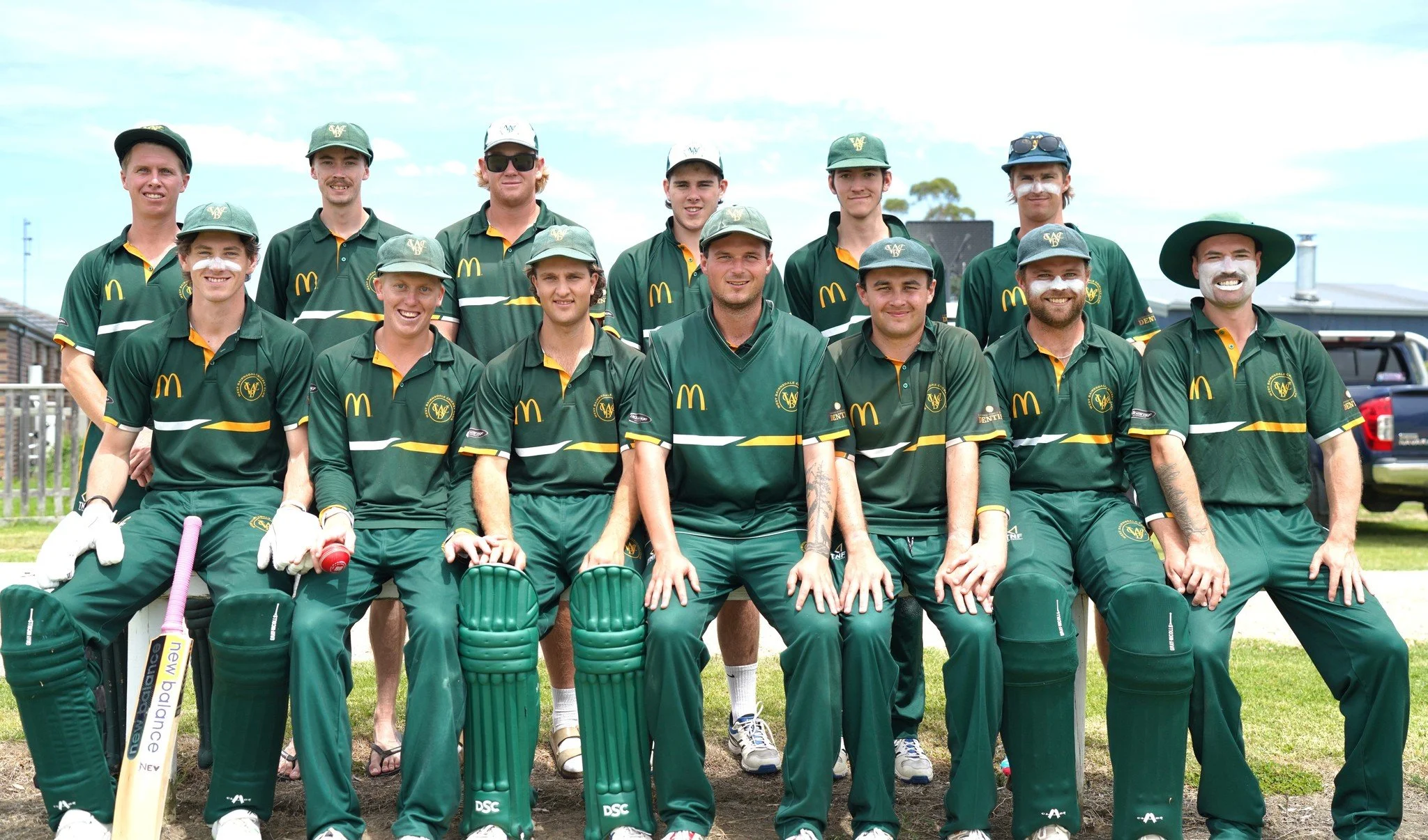 A group of fourteen men dressed in emerald green cricket uniforms, some wearing caps and others hats, posing outdoors on a grassy area under a partly cloudy sky. They are standing and sitting in two rows, with cricket equipment such as bats and pads visible. They are smiling and looking at the camera.