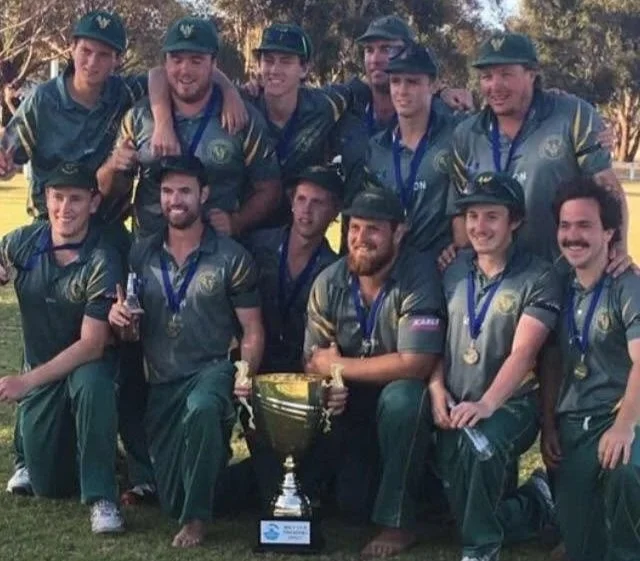 Cricket team wearing green uniforms and hats, posing outdoors with a trophy and medals, celebrating their victory.