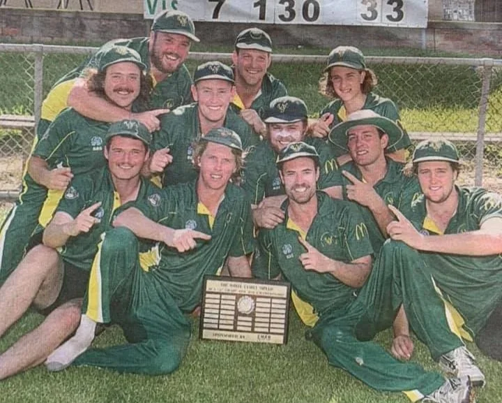 Group of young men in green and yellow sports uniforms celebrating on a baseball field, holding a plaque, with a scoreboard in the background.